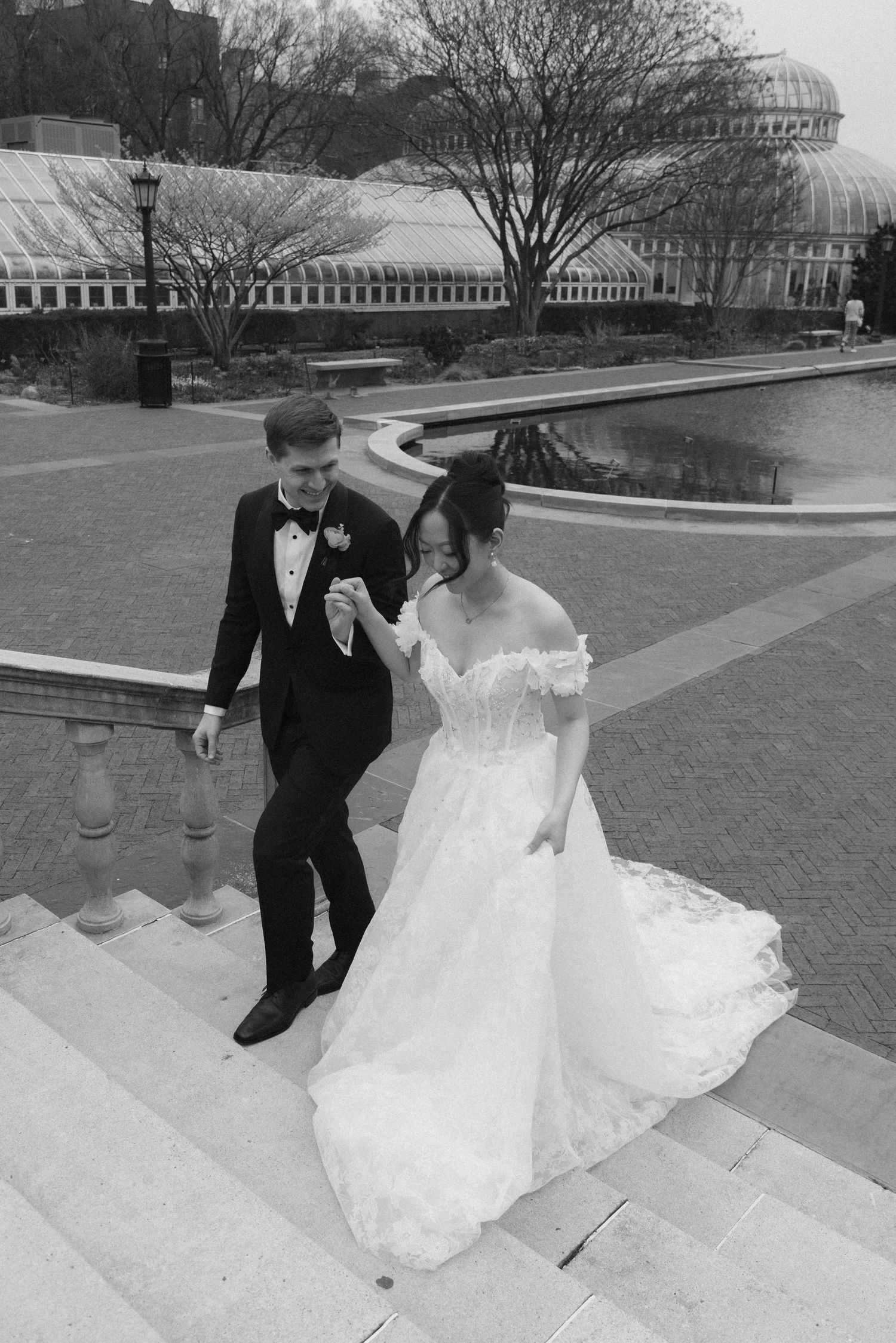 A couple descends stone steps outdoors on their wedding day, with the bride wearing a flowing white gown and groom in a tuxedo.