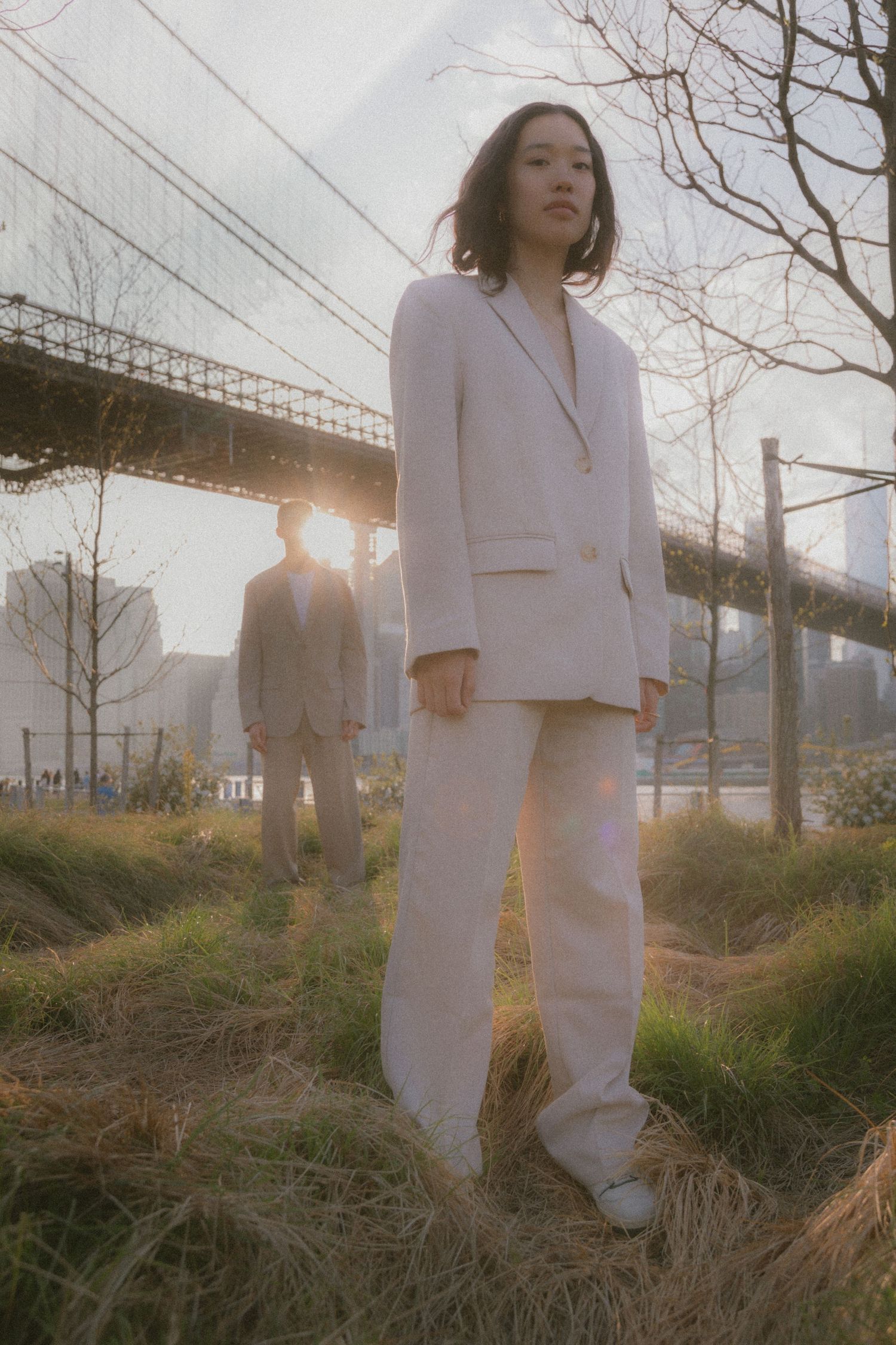 Two people in beige business suits pose on grassy terrain with a bridge silhouetted against a sunset sky.