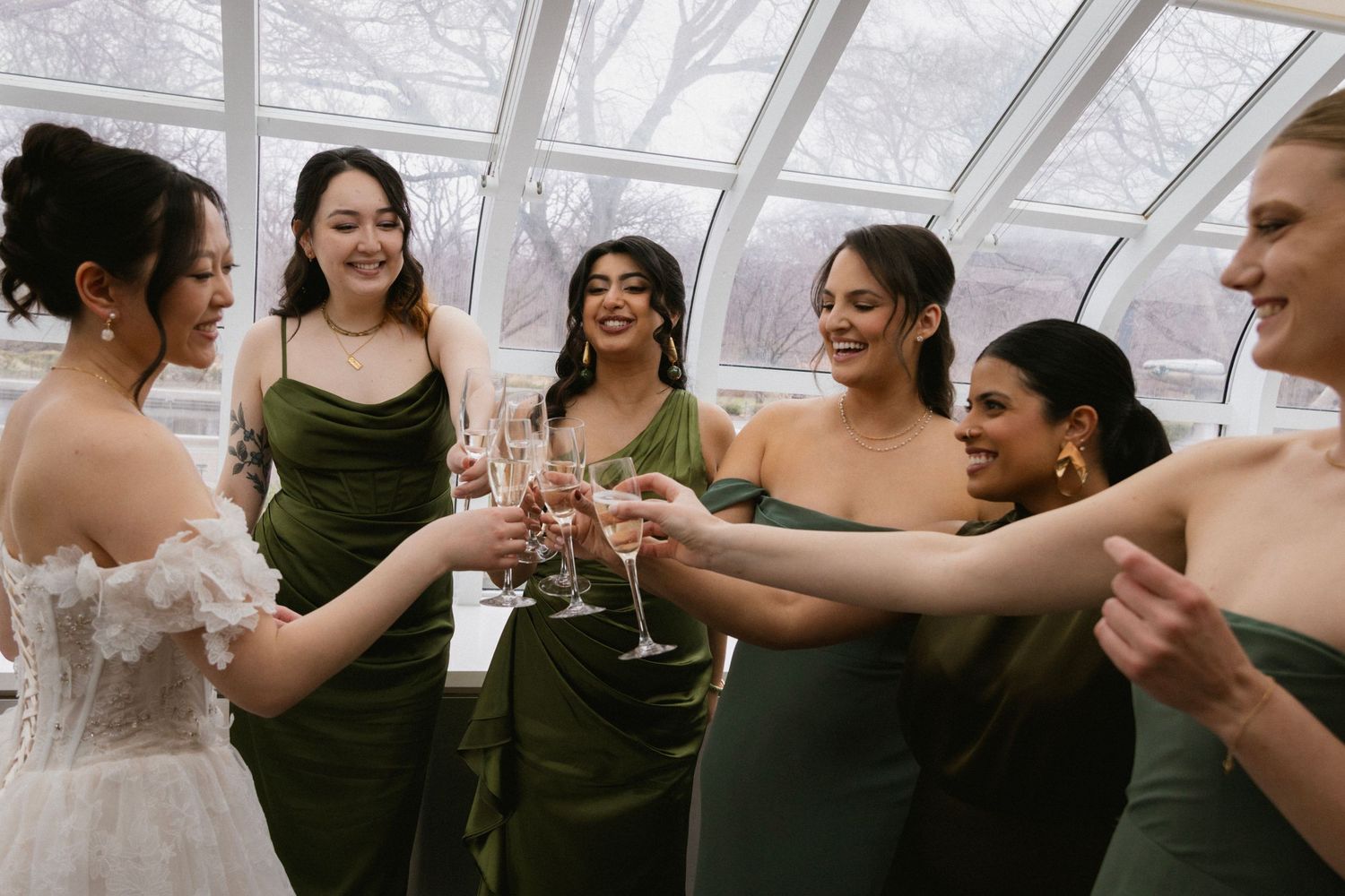 Bridesmaids in dark green dresses toast with champagne glasses in a sunlit greenhouse.