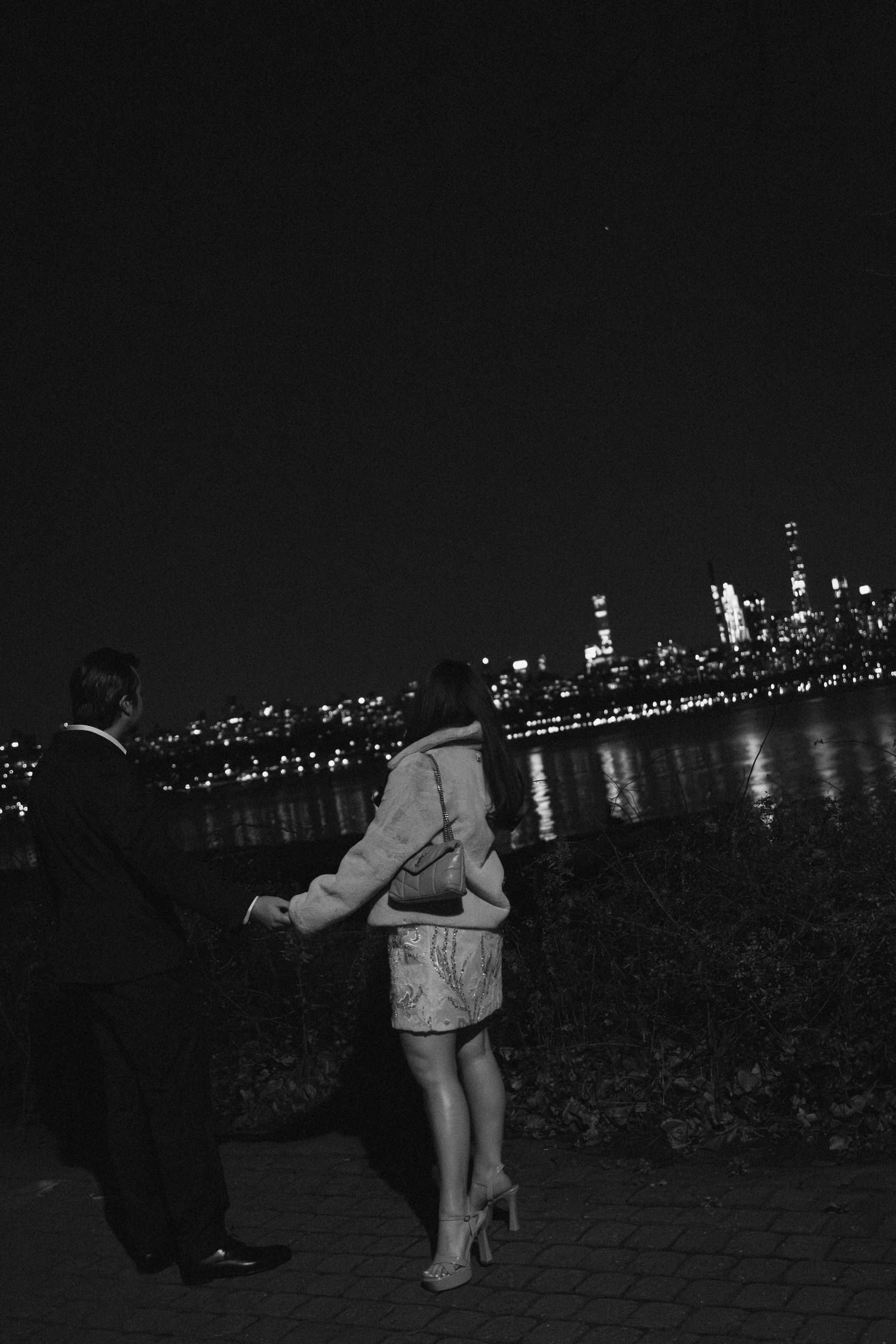 Two people stand along a waterfront path at night with an illuminated city skyline reflecting on the water.