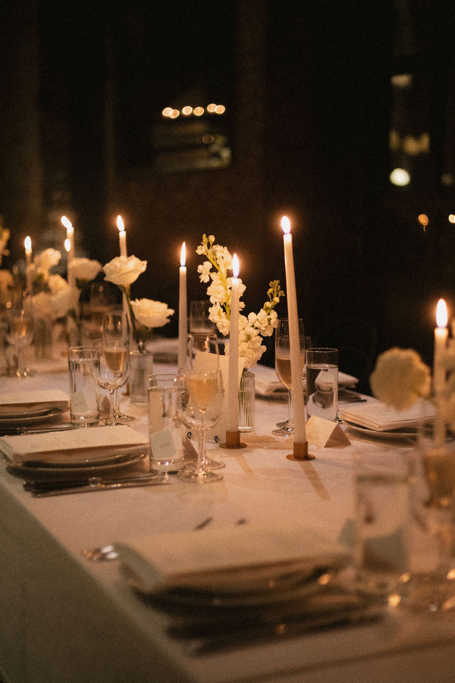 Elegant table setting with lit candles, white flowers in vases, and formal place settings for an intimate evening event.
