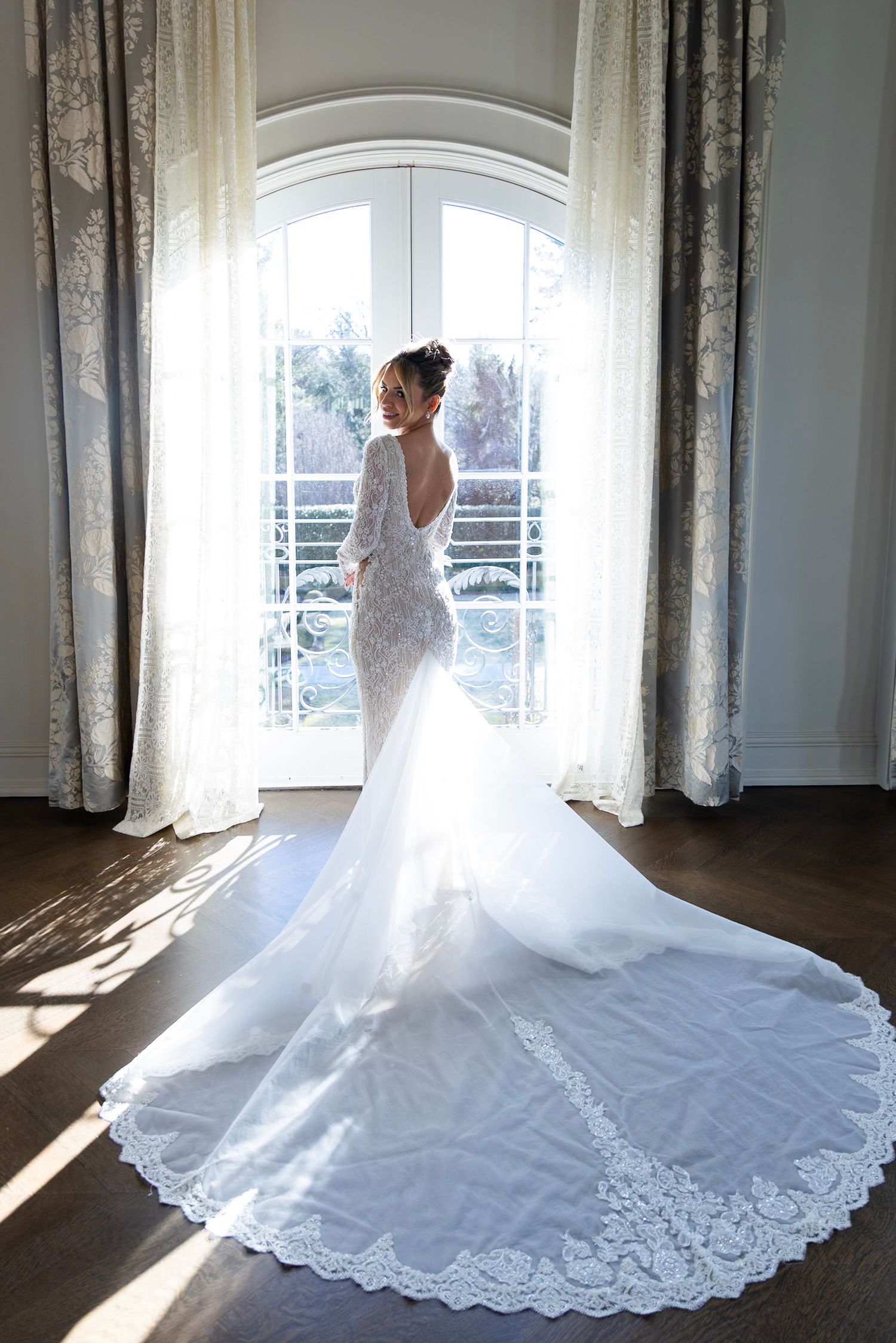 A bride in a fitted lace wedding gown with dramatic train poses by window in elegant room with flowing curtains.