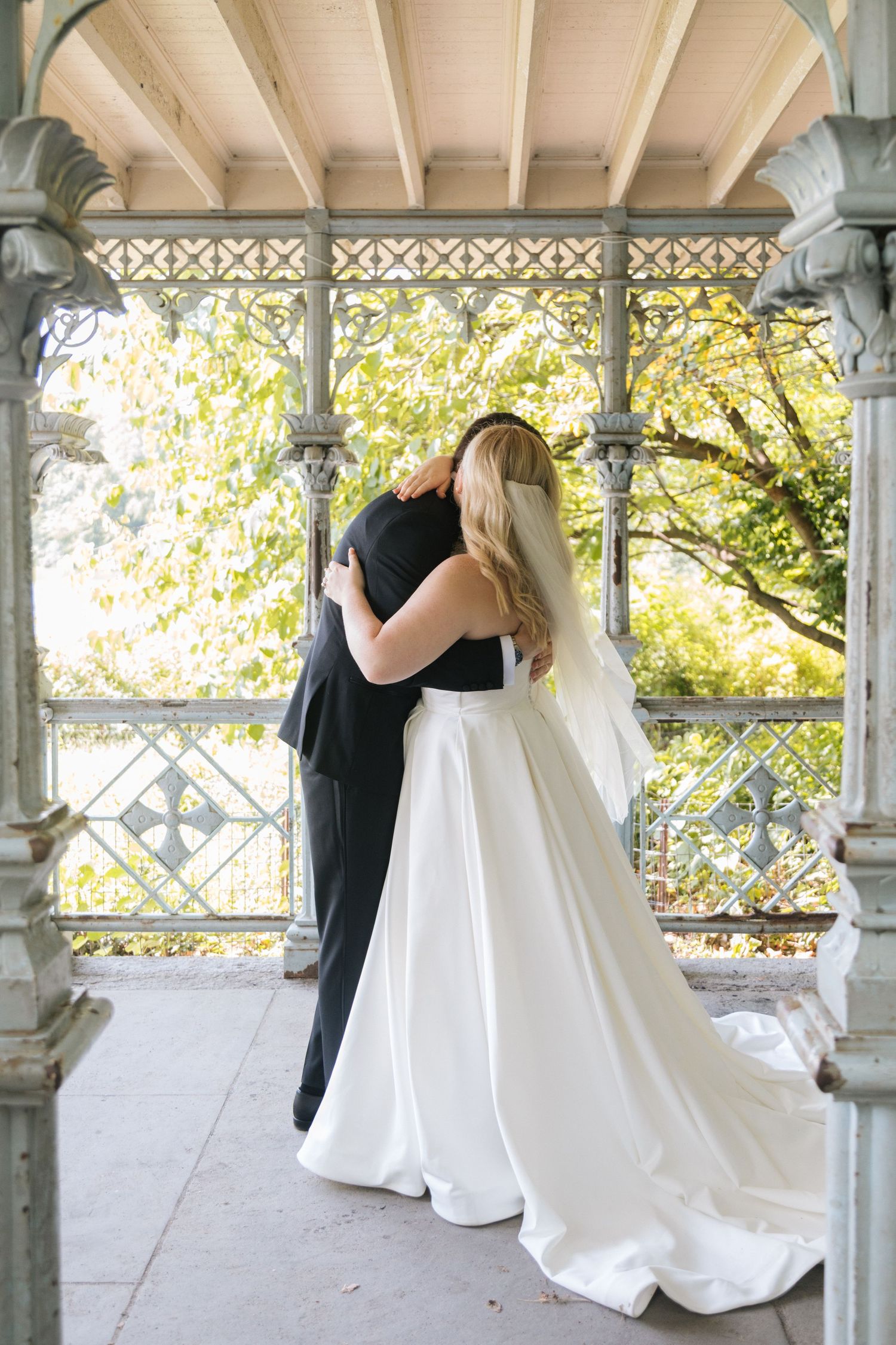 A couple in formal wedding attire embrace on an ornate Victorian-style porch with autumn foliage in the background.
