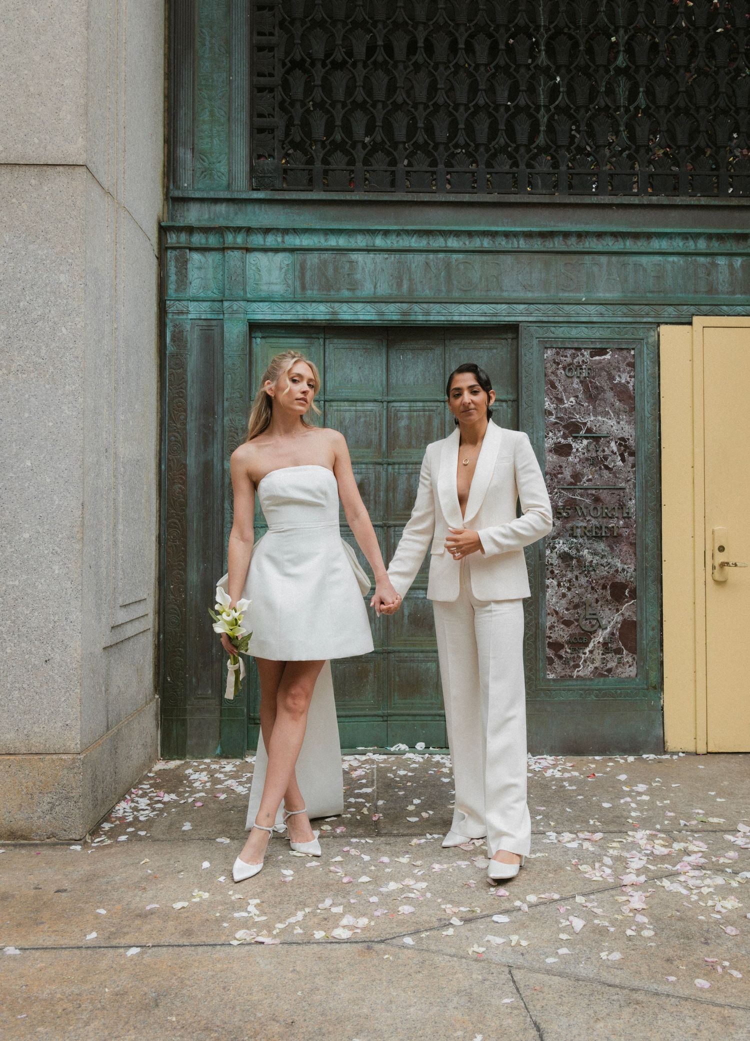 Two people in white outfits stand on a sidewalk covered with scattered white flower petals.