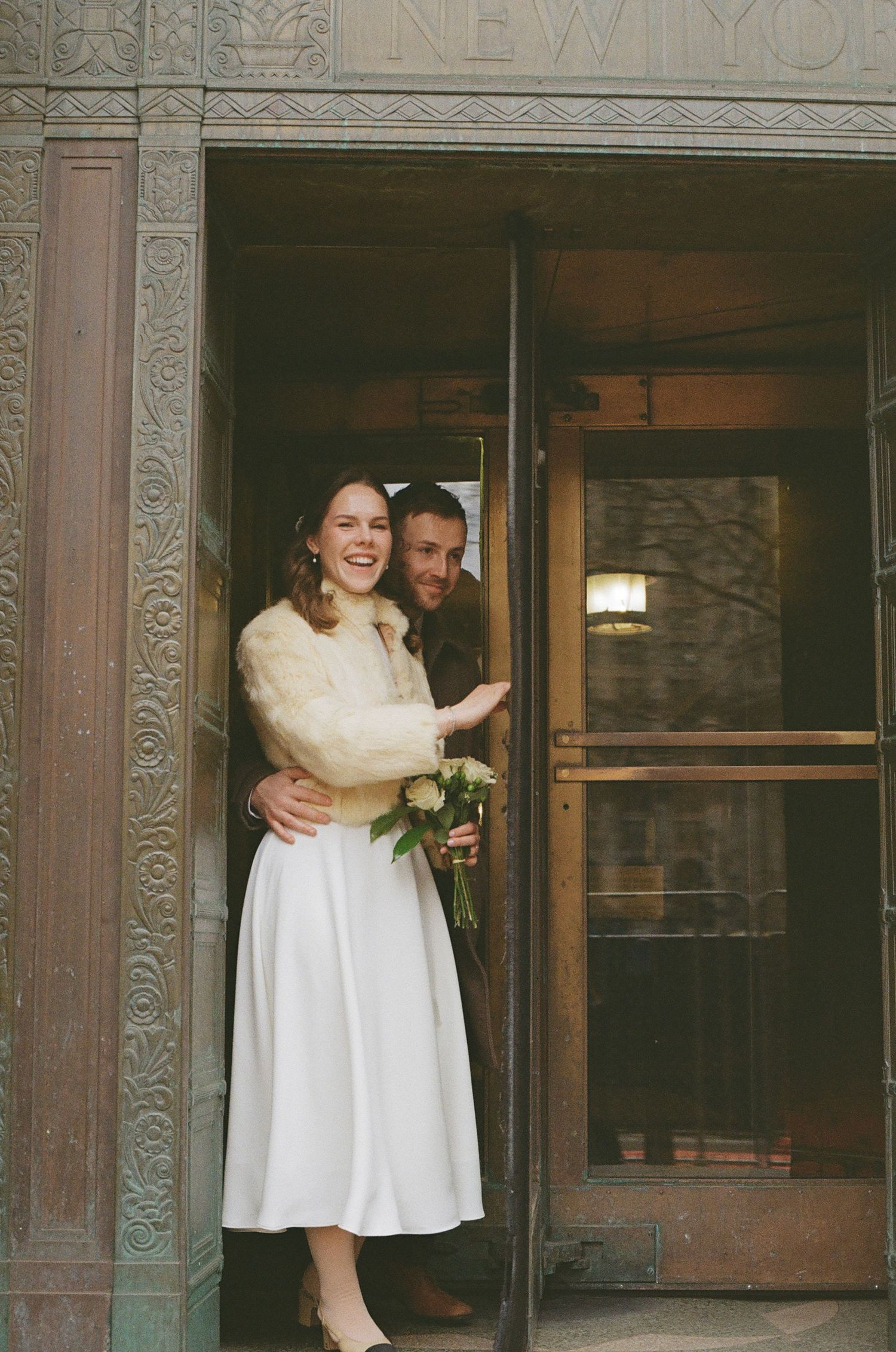 Bride and groom laughing in doorway of New York building, bride holding bouquet in white dress and fur shrug.