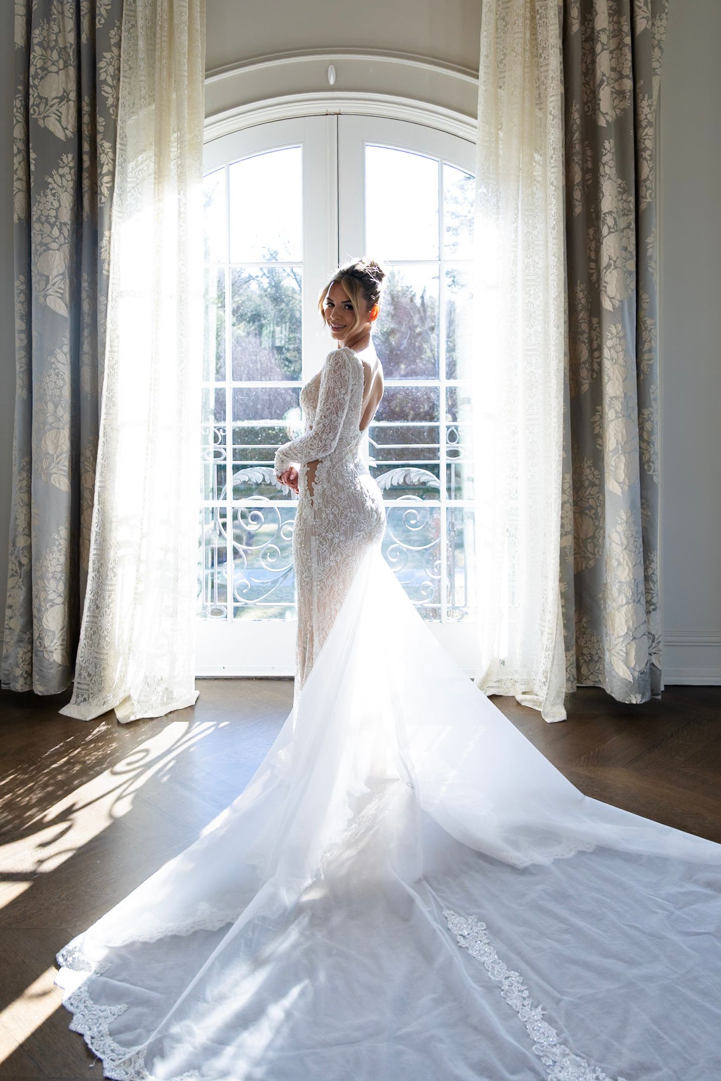 Beautiful lace wedding gown with long train photographed in natural light by arched window with flowing curtains.