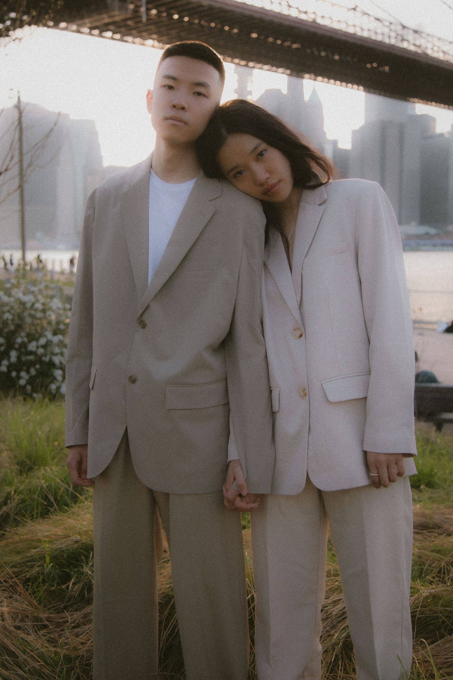 A couple in matching neutral-toned suits poses together outdoors under a bridge in a romantic photoshoot.