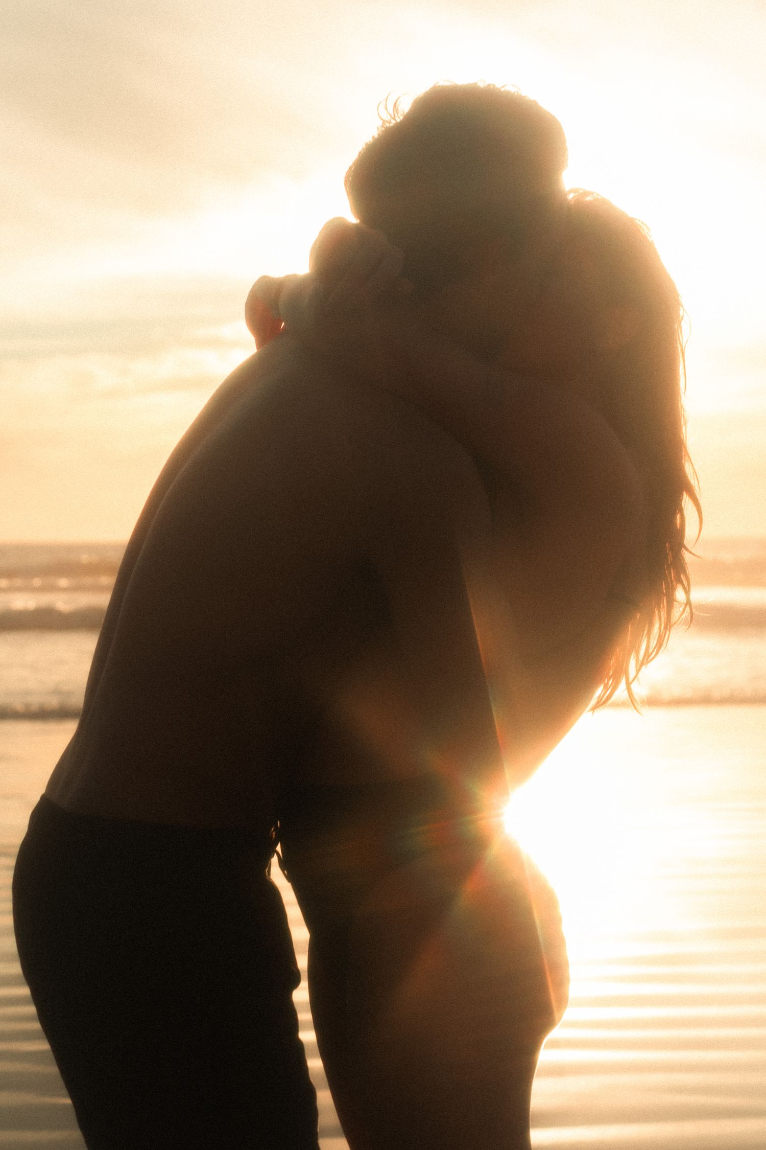 A silhouetted couple embraces on a beach at sunset with golden light streaming through their romantic pose.