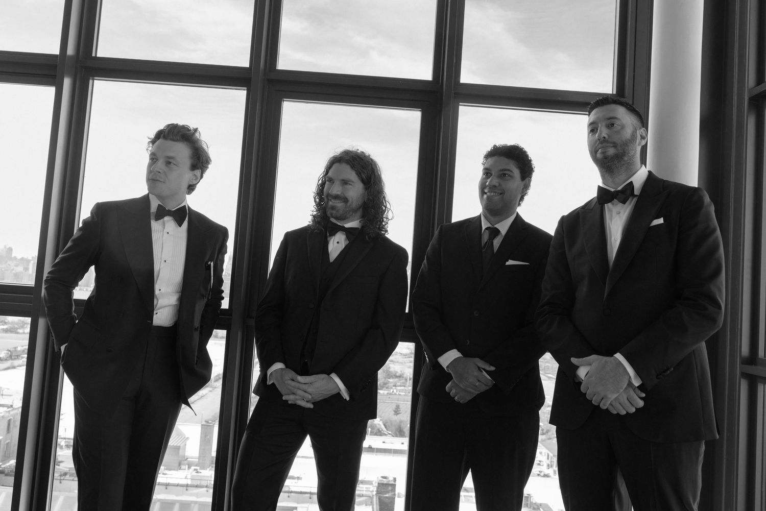 A wedding party in formal black attire stands in silhouette against bright windows in an elegant venue.