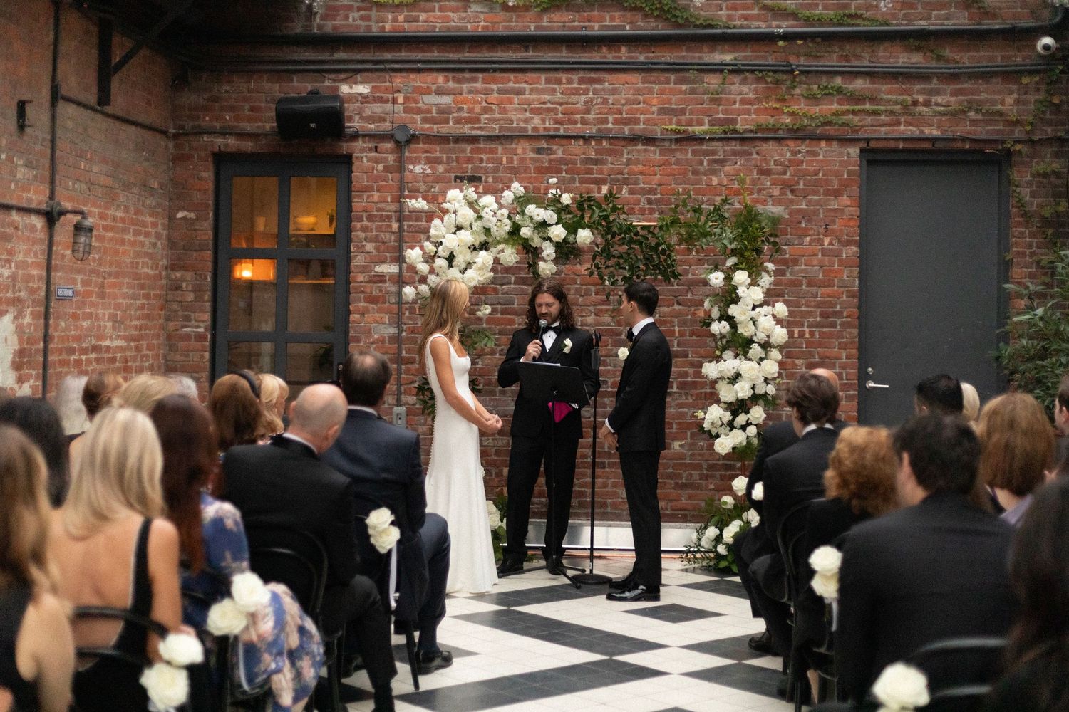 Wedding ceremony guests gather in an outdoor courtyard surrounded by historic brick buildings.