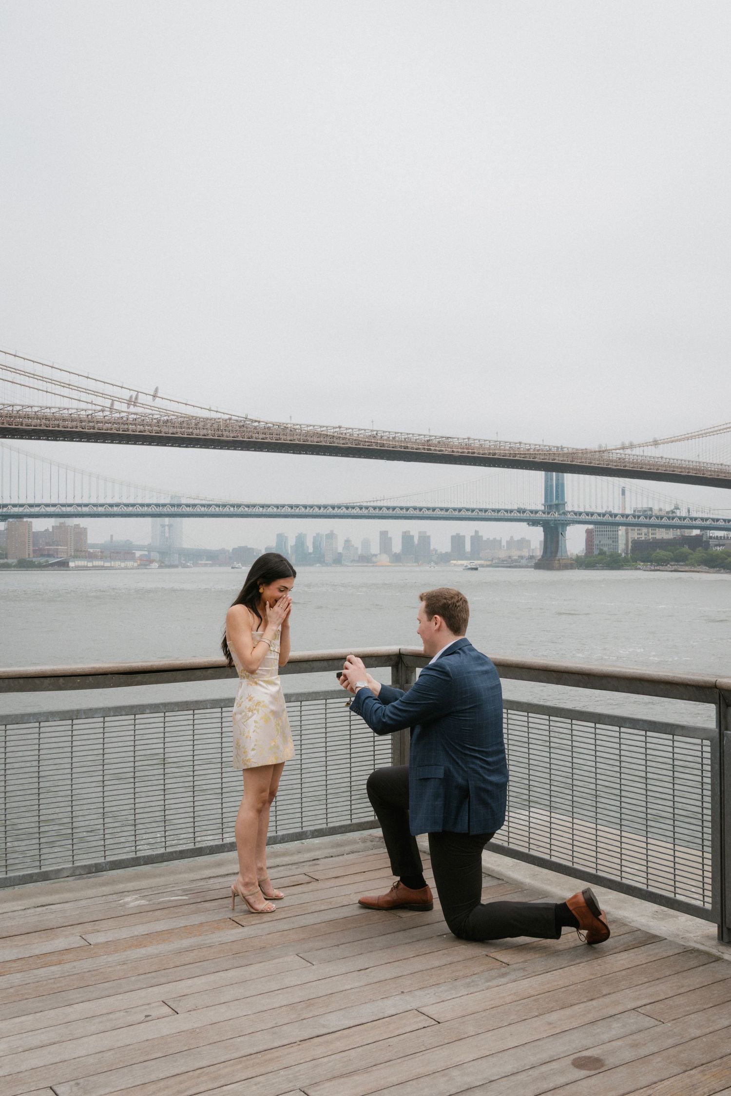 Romantic proposal scene on waterfront deck with Brooklyn Bridge in background.