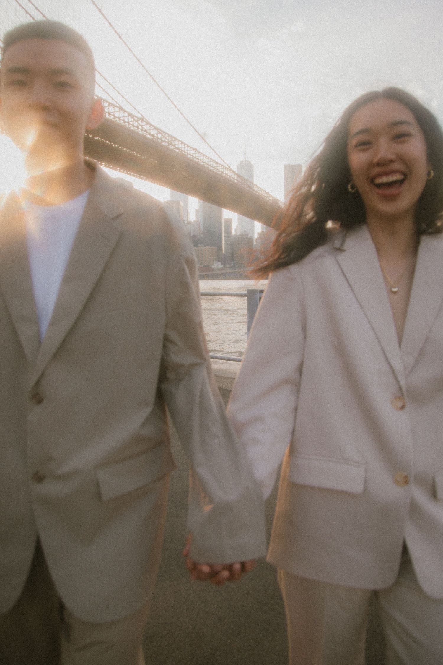 Happy couple in matching light suits hold hands at sunset with Brooklyn Bridge in background.