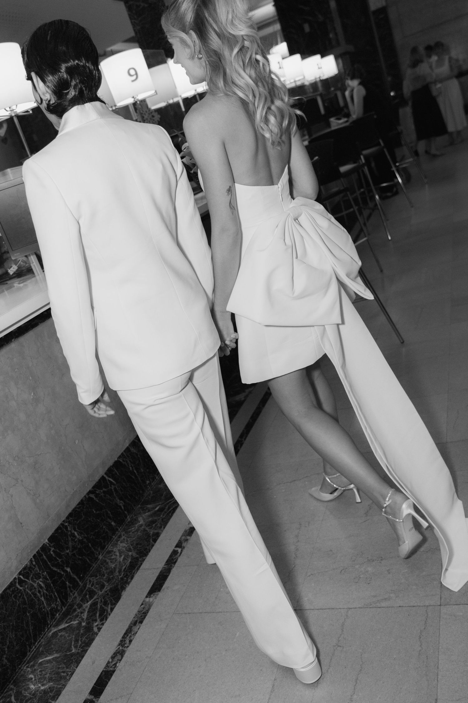 A black and white photo showing a couple in white formal attire walking together.