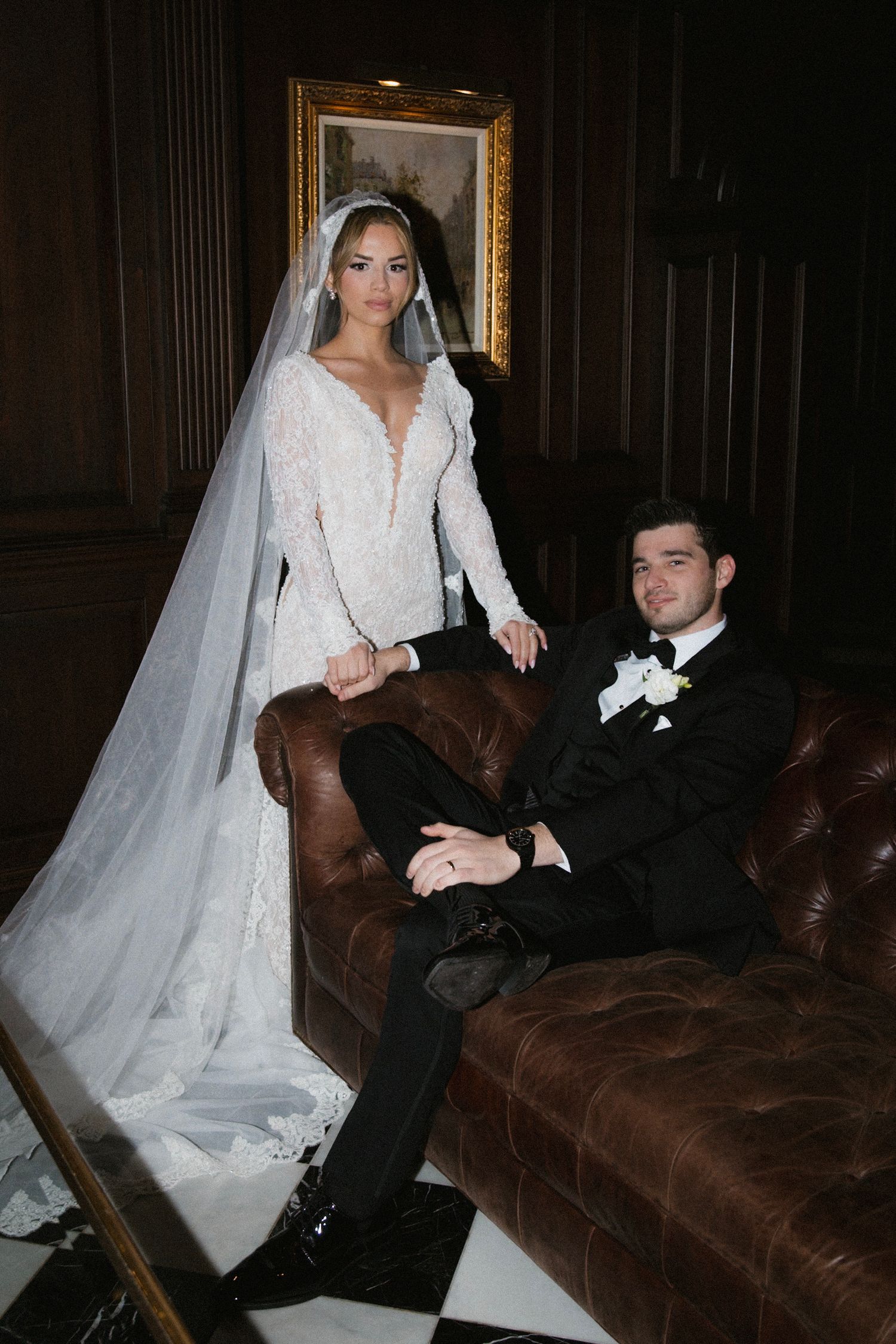 Wedding photoshoot on a brown leather couch shows a bride in white dress and veil posing with groom in dark suit.
