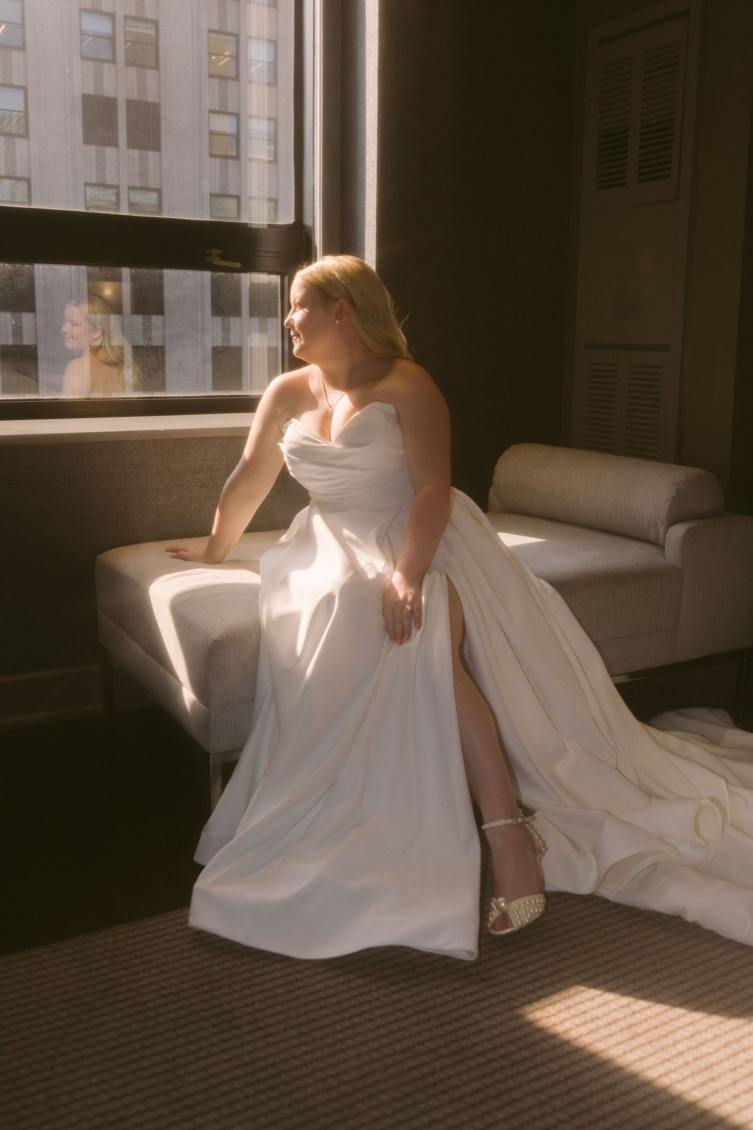 Bride in white wedding dress sitting elegantly on couch with sunlight streaming through nearby window.