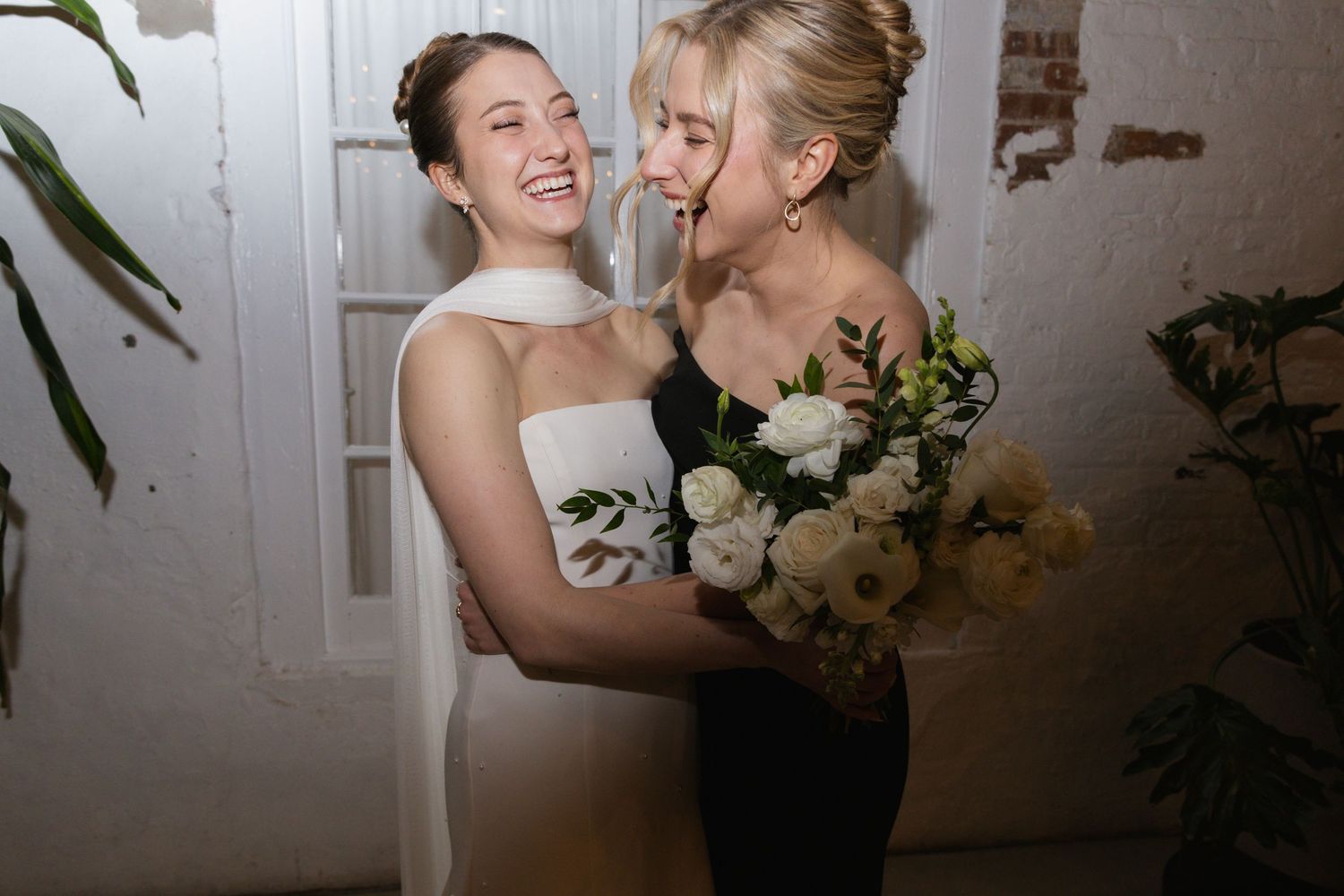 Two wedding guests share a joyful moment with white flowers against a white wall.