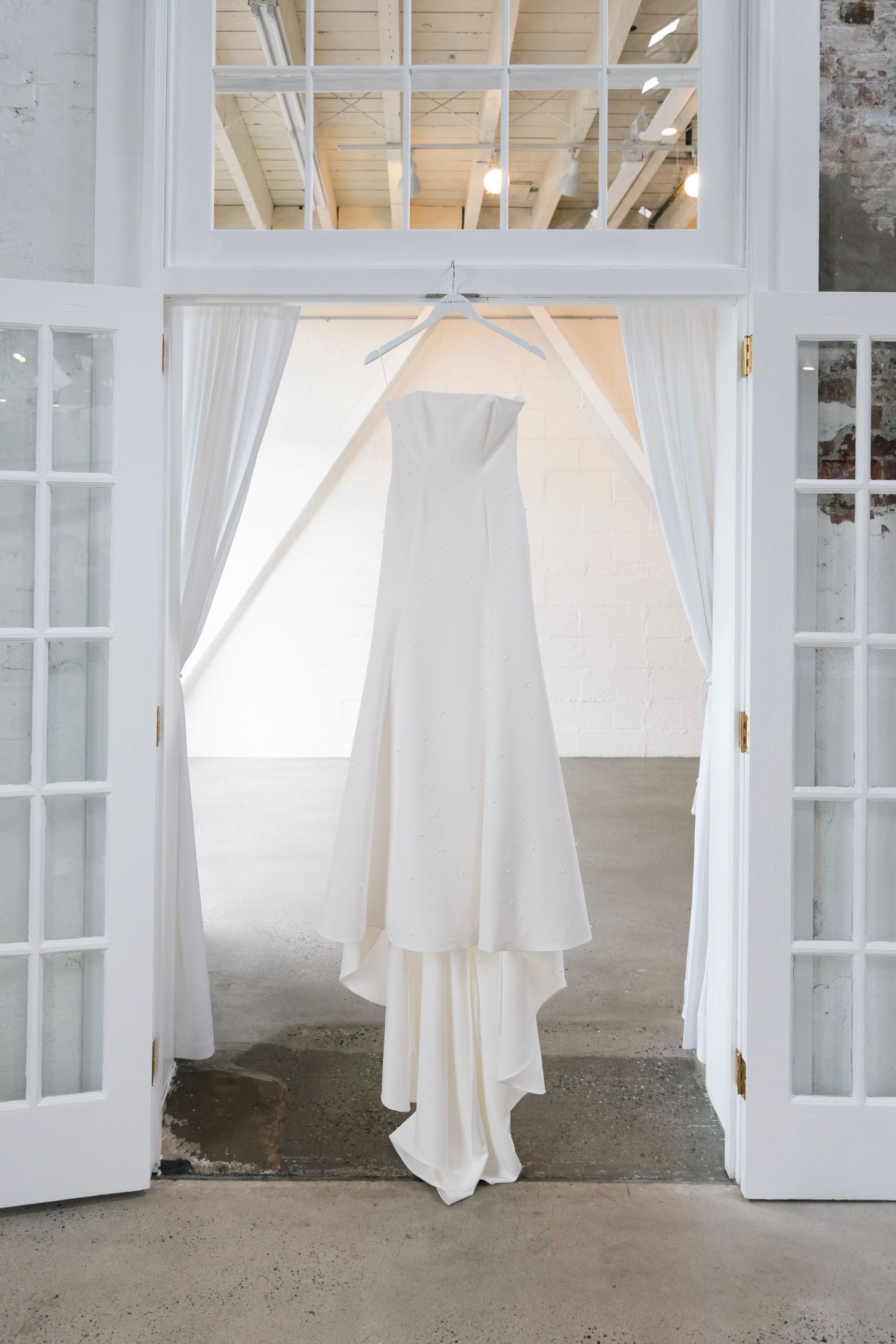 A white wedding dress hangs elegantly in a doorway framed by French doors and flowing sheer curtains.