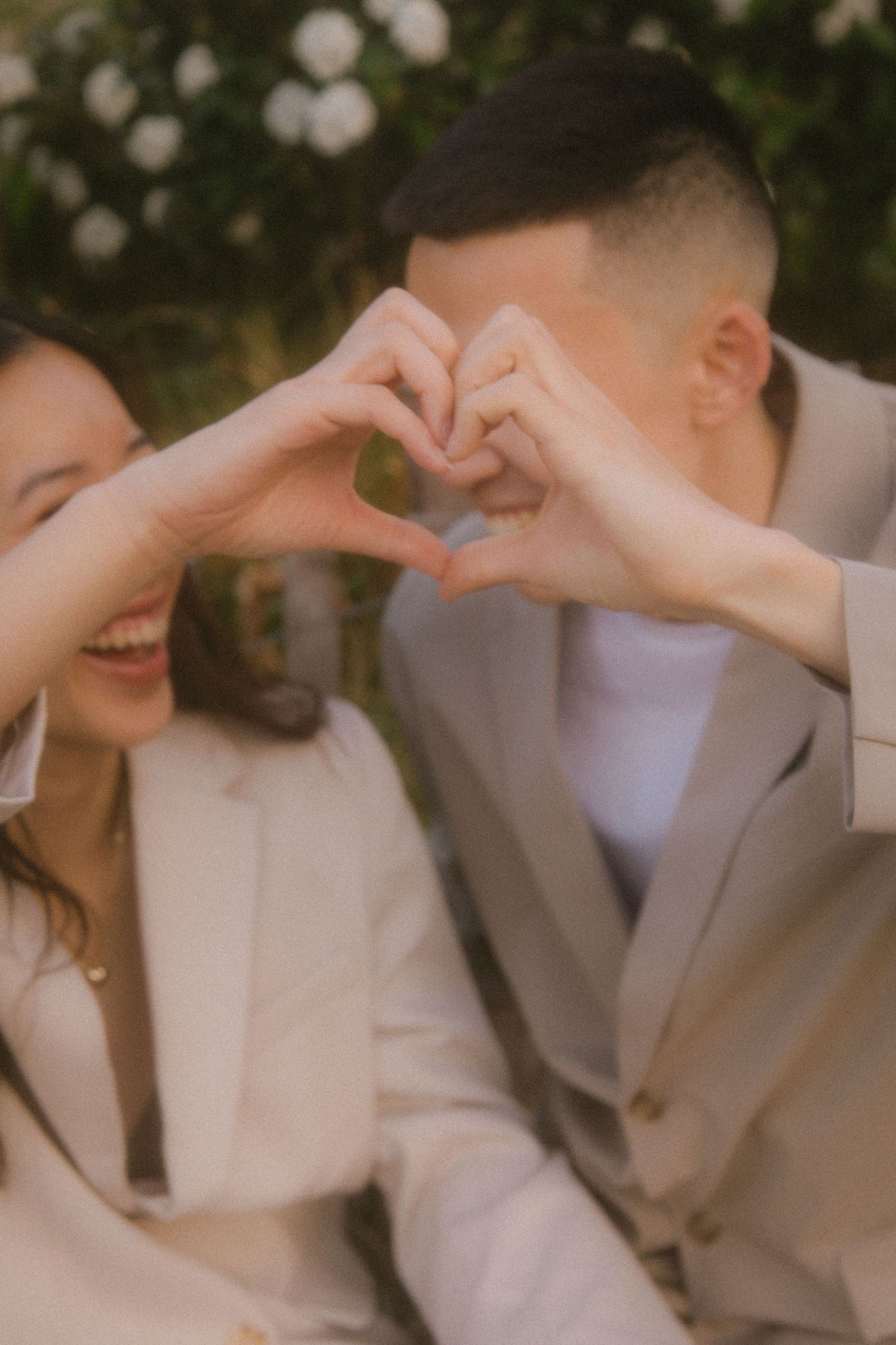 Two people in matching light colored blazers make a heart shape with their hands while smiling.