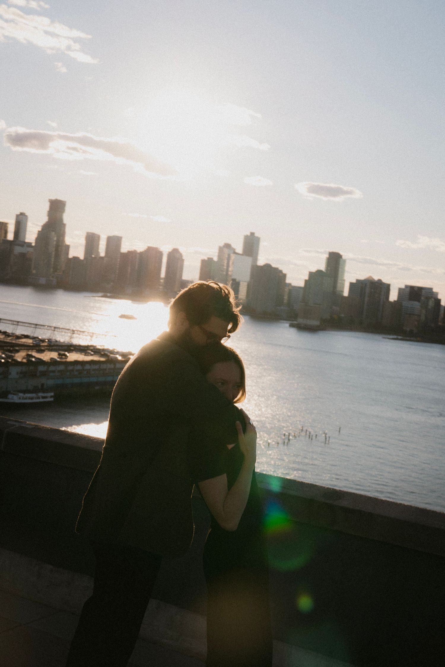 A silhouetted couple embraces while overlooking a city skyline at sunset with lens flare and water reflections.