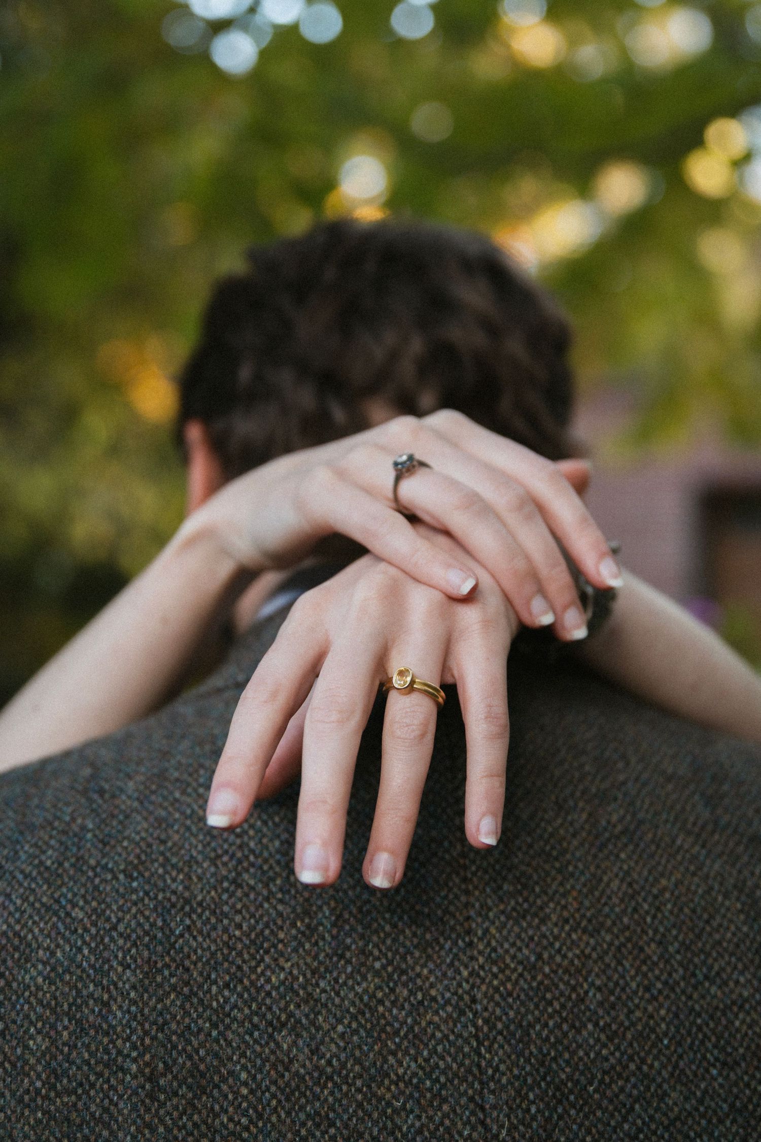 Close-up of hands with engagement rings embracing over a shoulder against blurred natural background.