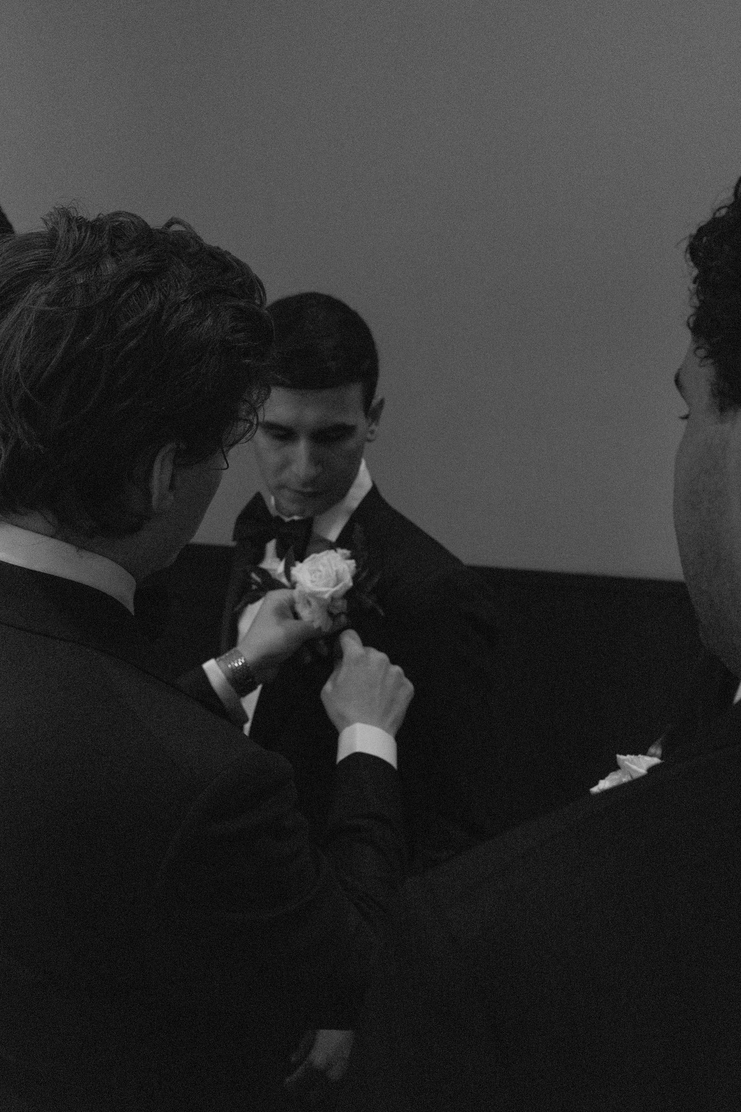 Groomsmen help pin a white boutonniere flower on a suit jacket in this black and white wedding moment.
