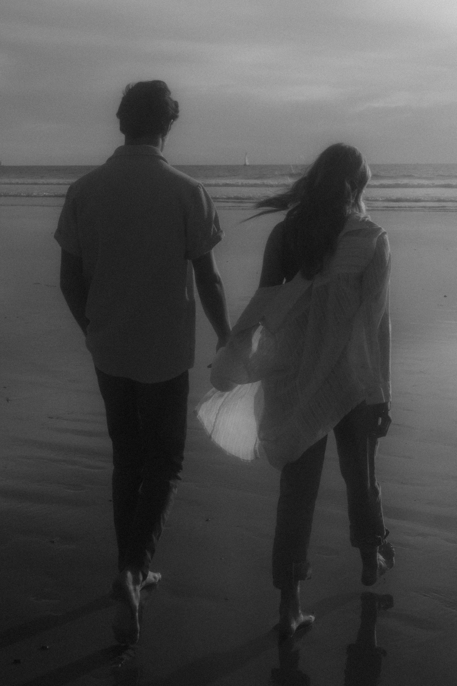 Two silhouettes walk together on a misty beach during sunset, their figures reflected in the wet sand below.