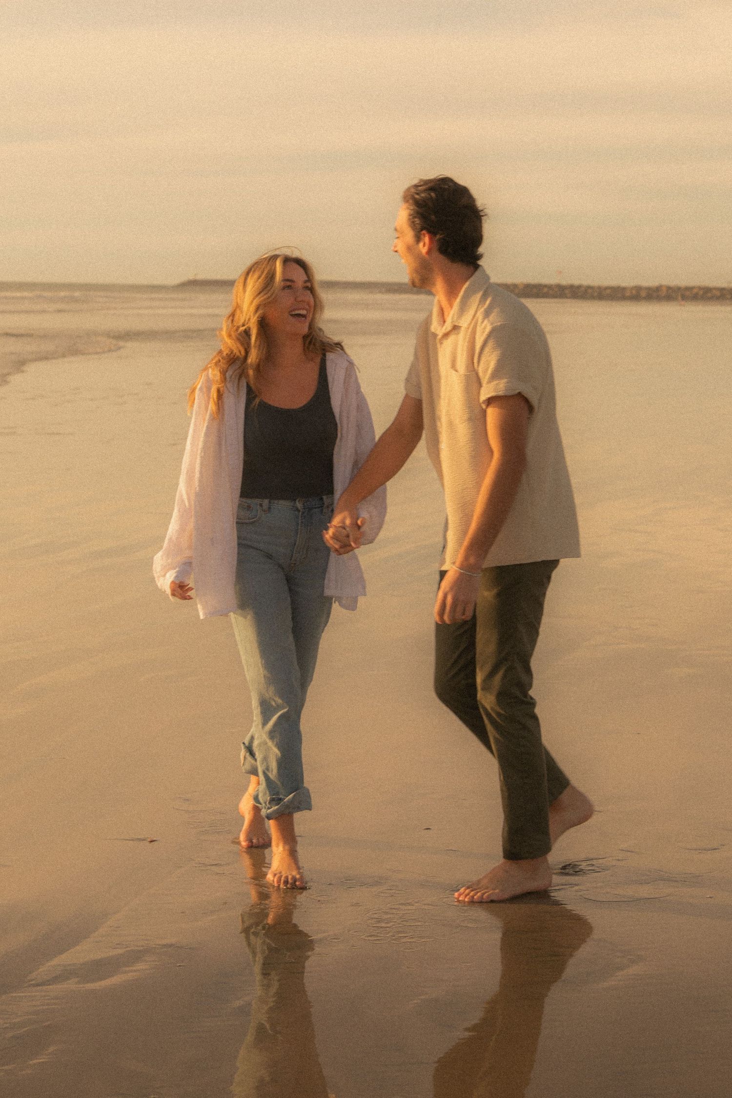A couple walks hand in hand on a sandy beach at sunset, leaving footprints in the wet sand.