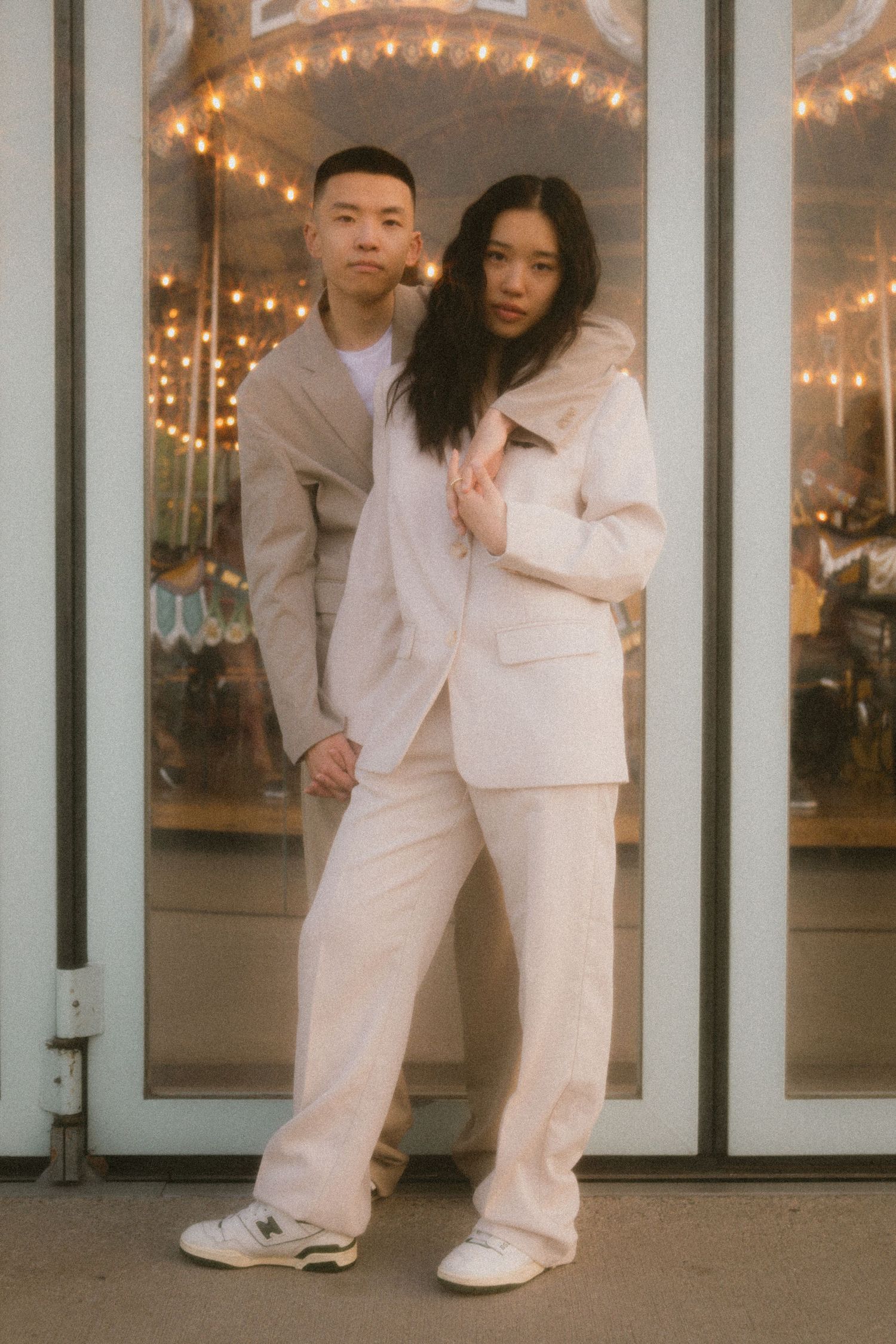 A couple in coordinated white outfits poses near a building with string lights, creating a romantic evening ambiance.