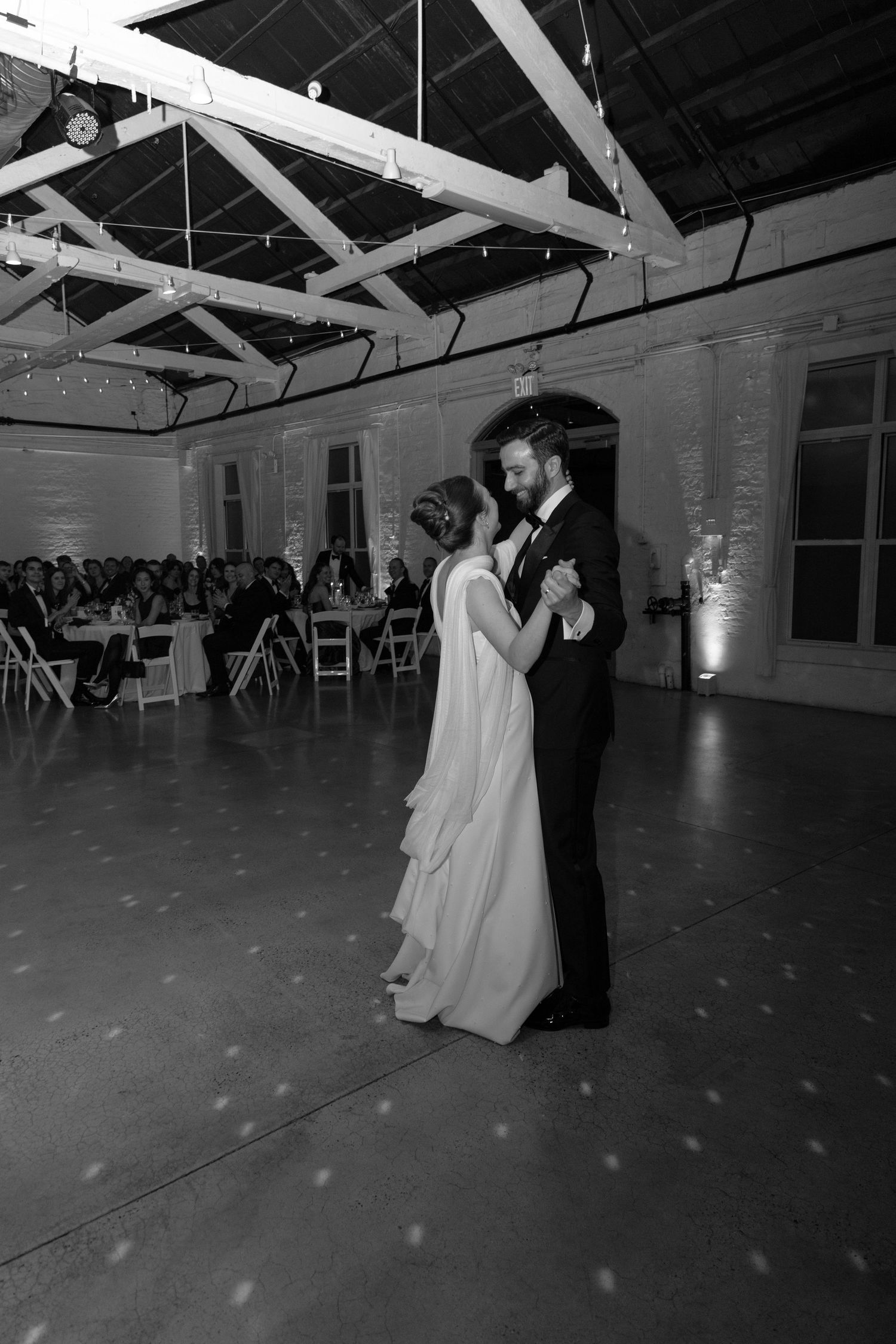 A romantic black and white photograph of a couple's first dance in an industrial-style reception venue.