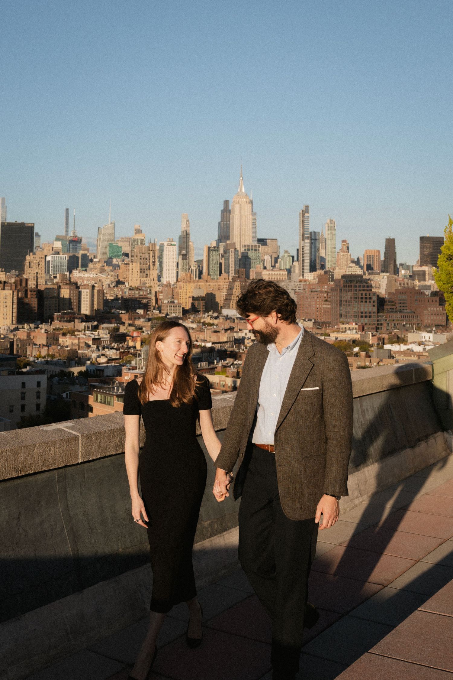 A couple stands together on a rooftop with the New York City skyline visible in the background during golden hour.