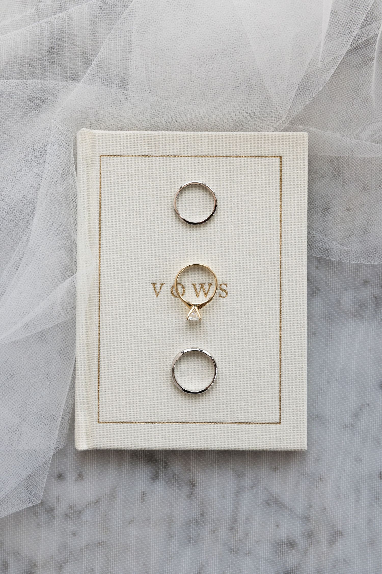 Wedding rings and vows book arranged on white marble surface with delicate tulle fabric.
