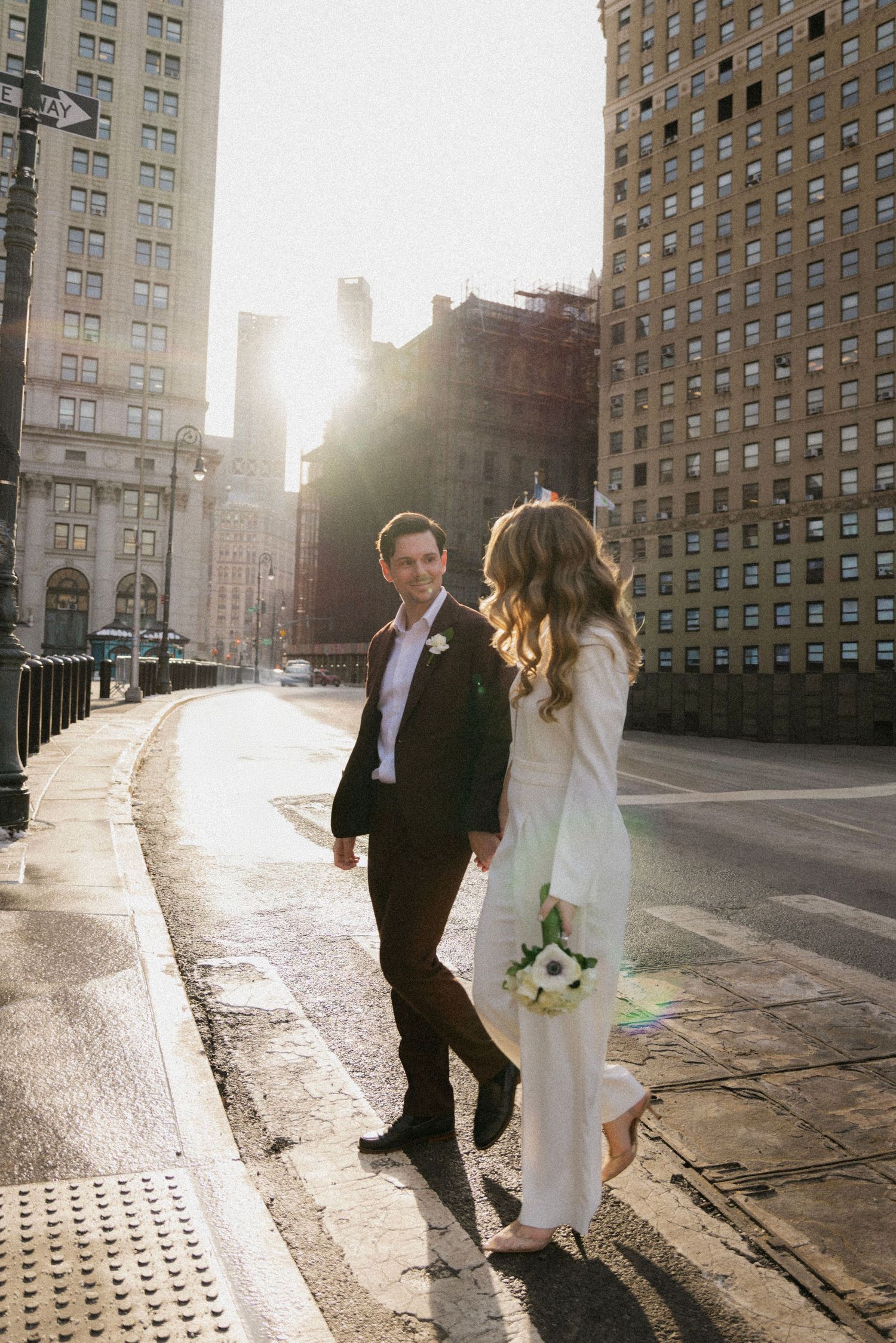 Bride and groom walk hand in hand on a sunlit city street, bride holding white bouquet in elegant white gown.