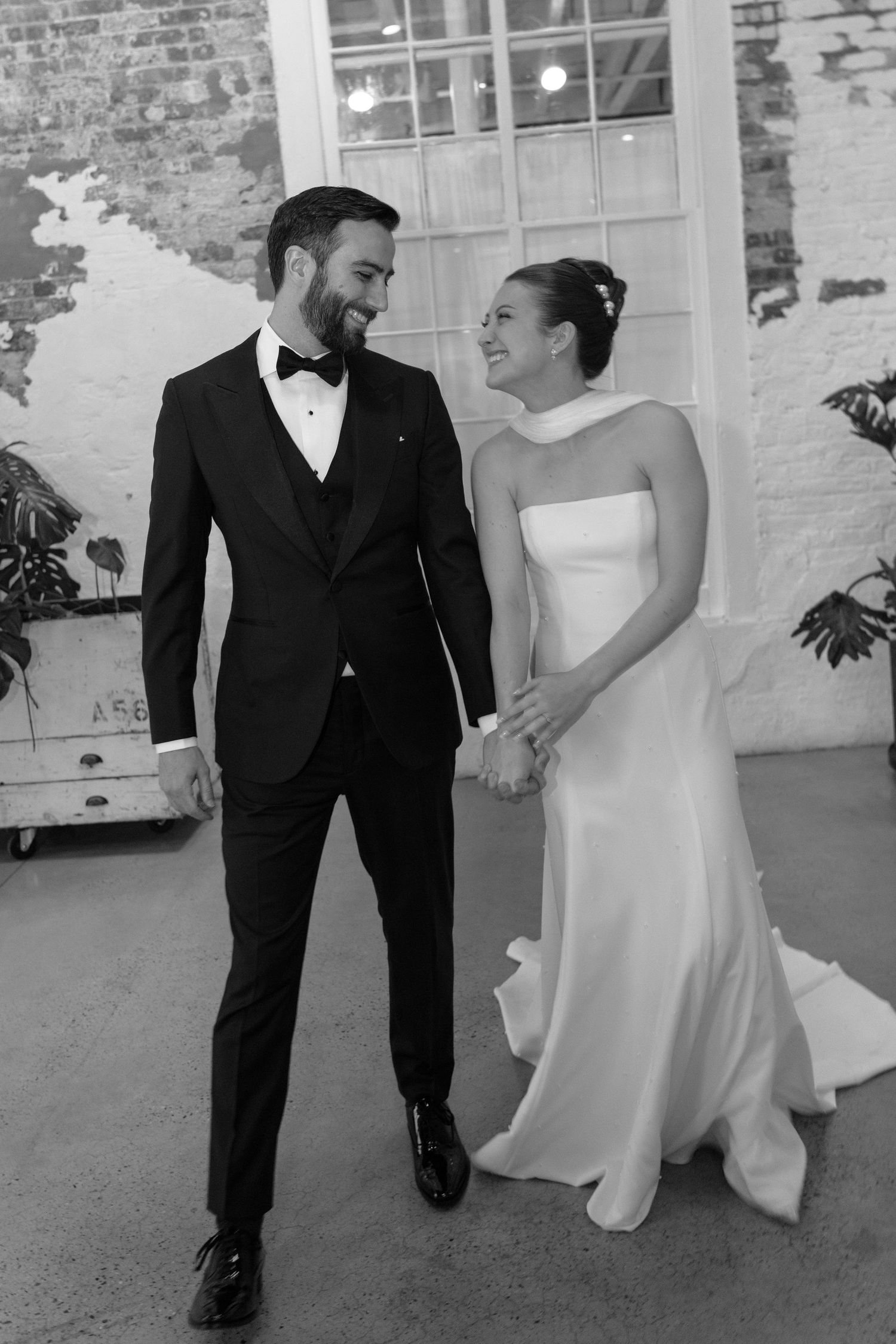 Black and white wedding photo showing a couple in formal attire sharing a joyful moment against a brick wall background.