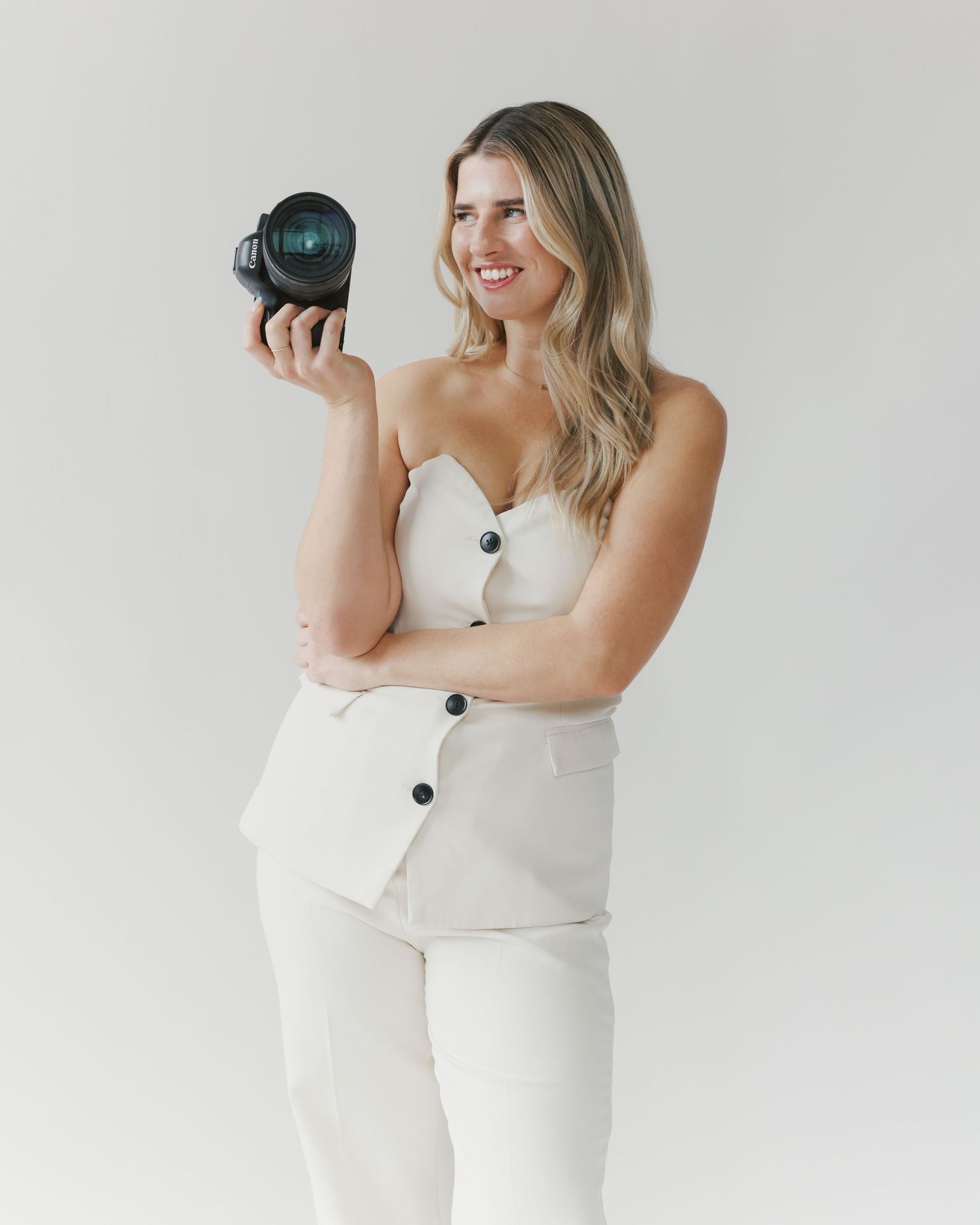 Professional portrait of woman in white outfit holding camera against light background.