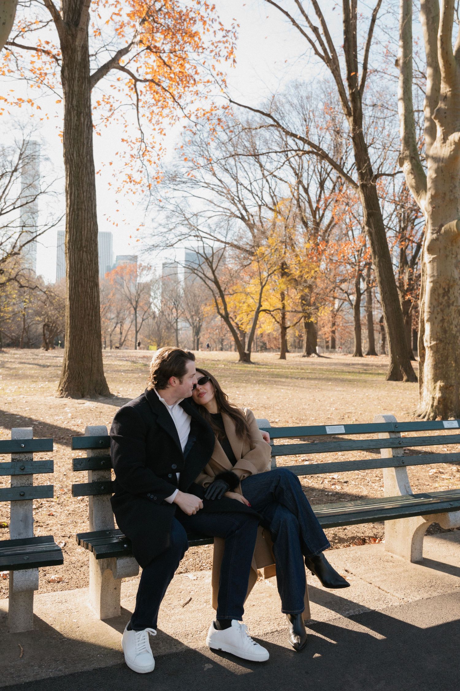 Two people sit close together on a wooden bench in a park during autumn with fallen leaves on the ground.