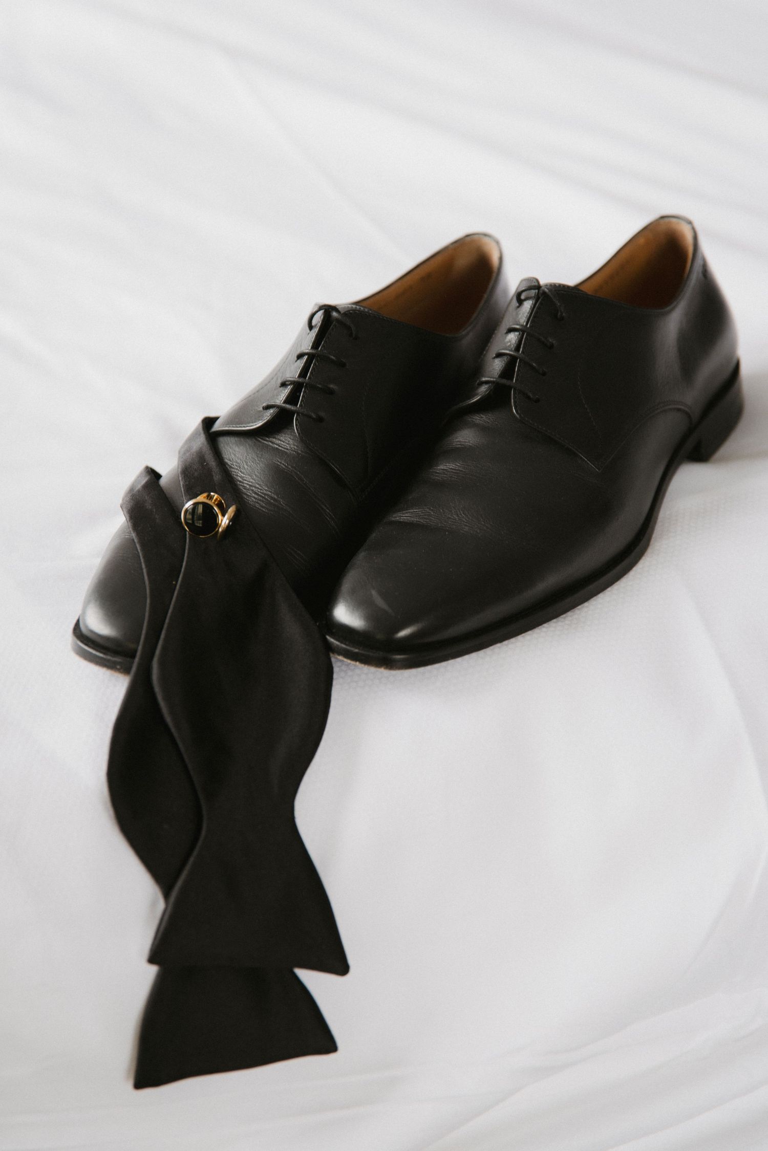 Classic black dress shoes and bow tie arranged on white fabric.