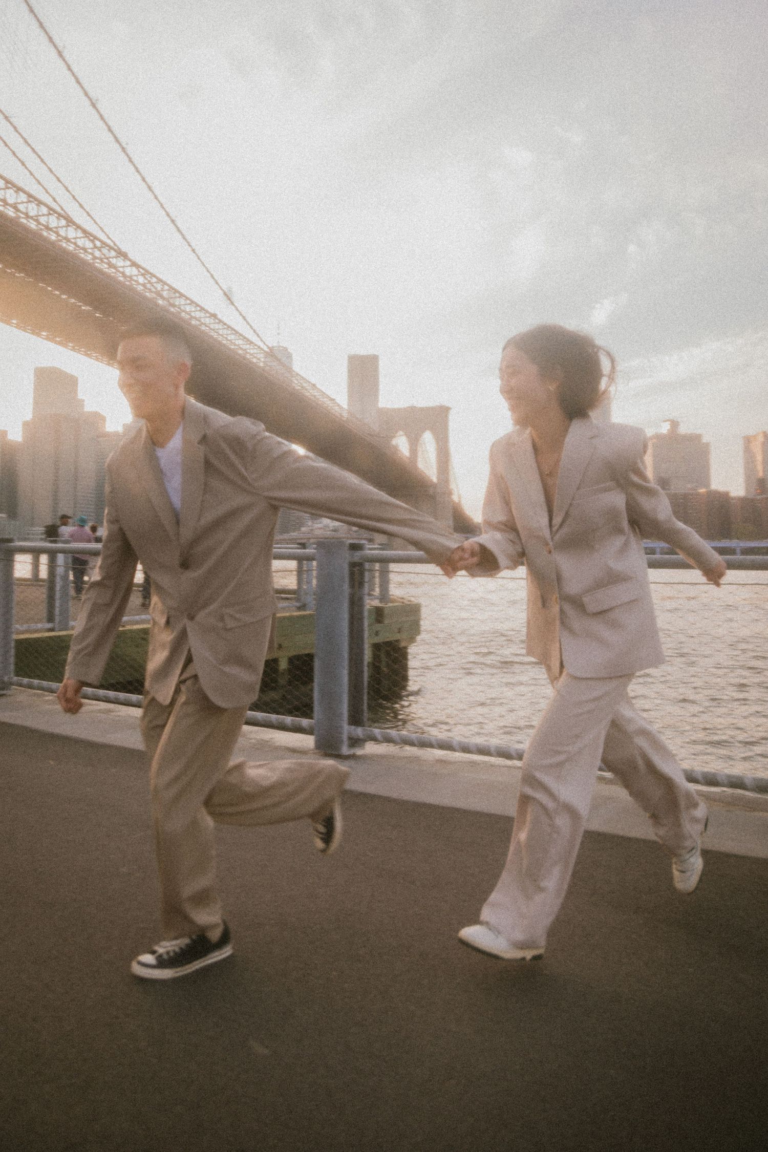 A couple in matching light suits playfully running along the waterfront with Brooklyn Bridge in the background at sunset.