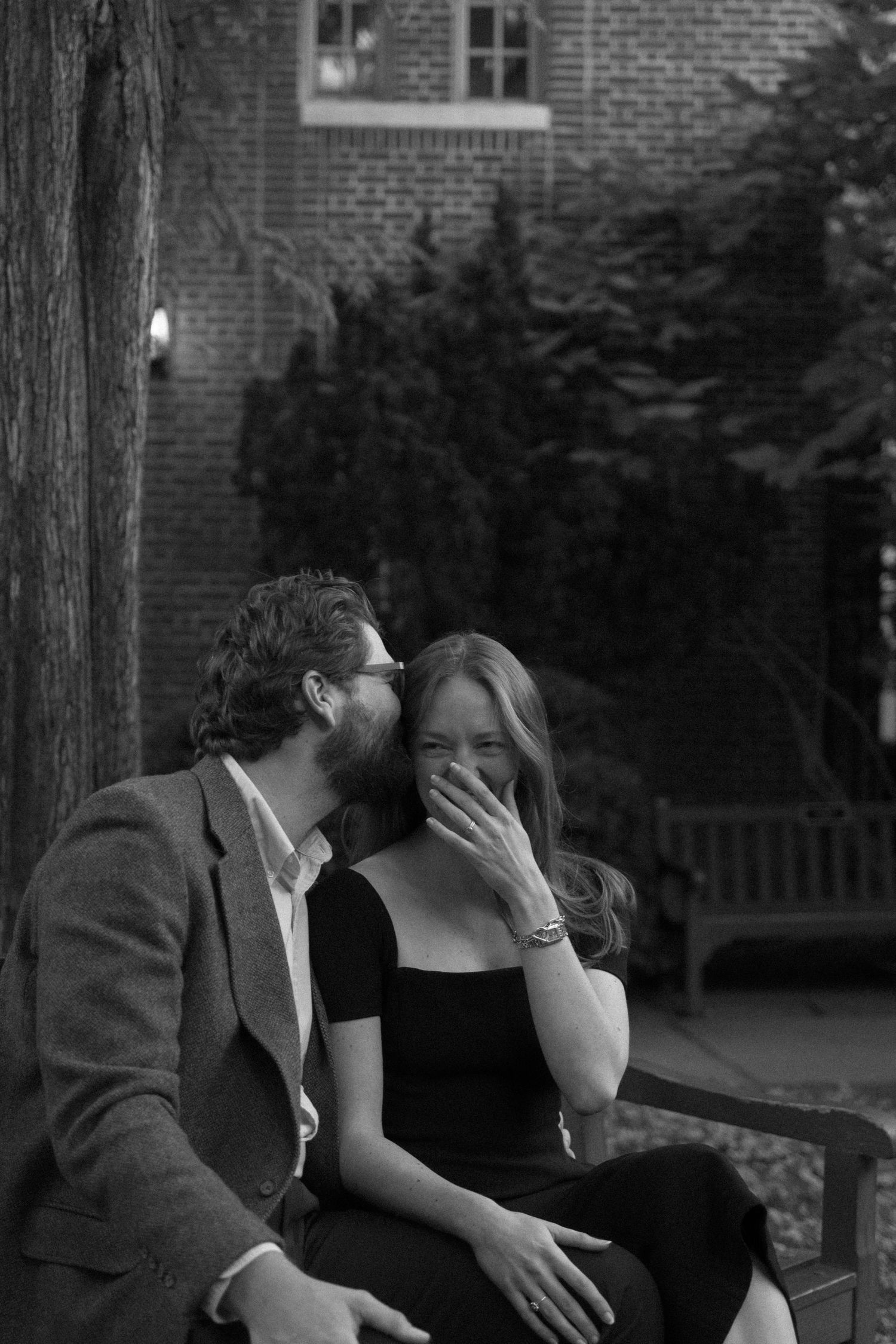 A couple in black and white share an intimate moment on a bench outside a brick building.