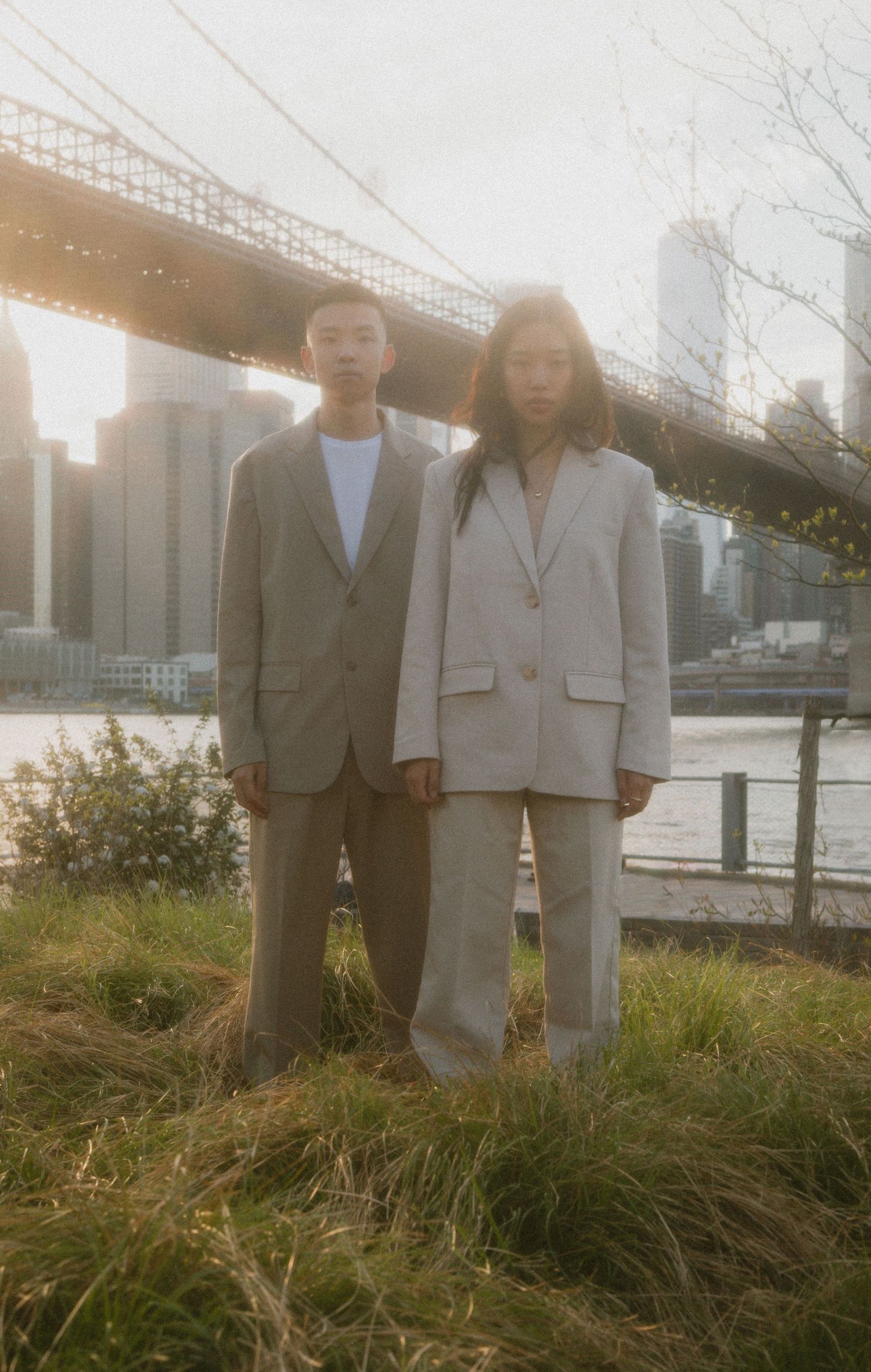 Two people in oversized suits stand on grass with the Brooklyn Bridge and NYC skyline in the background.