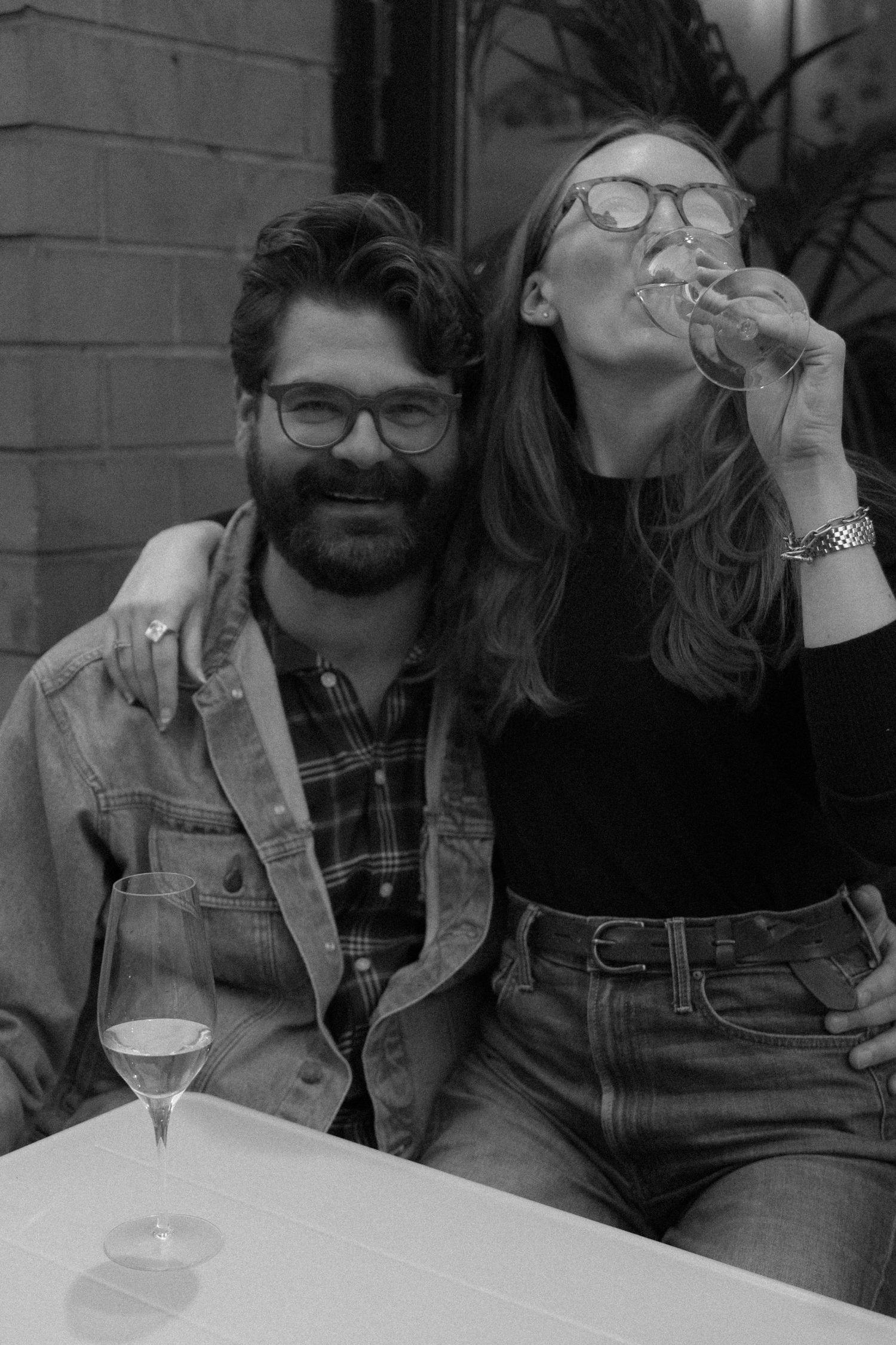 A couple laughs and shares drinks together at an outdoor table in black and white.