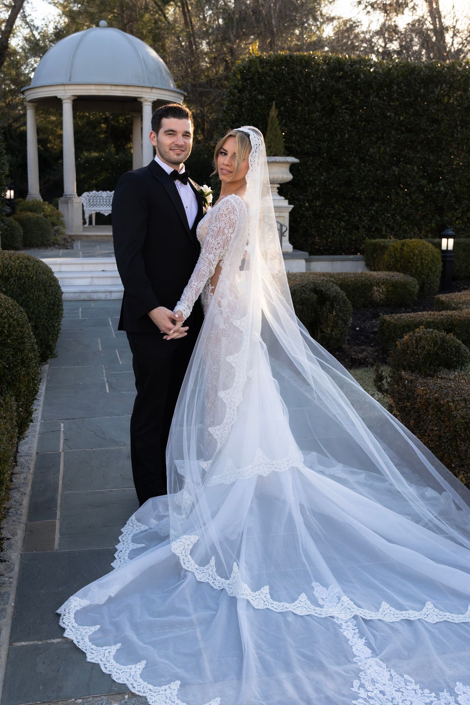 A stunning cathedral-length wedding veil trails behind on stone pavement near a garden gazebo.