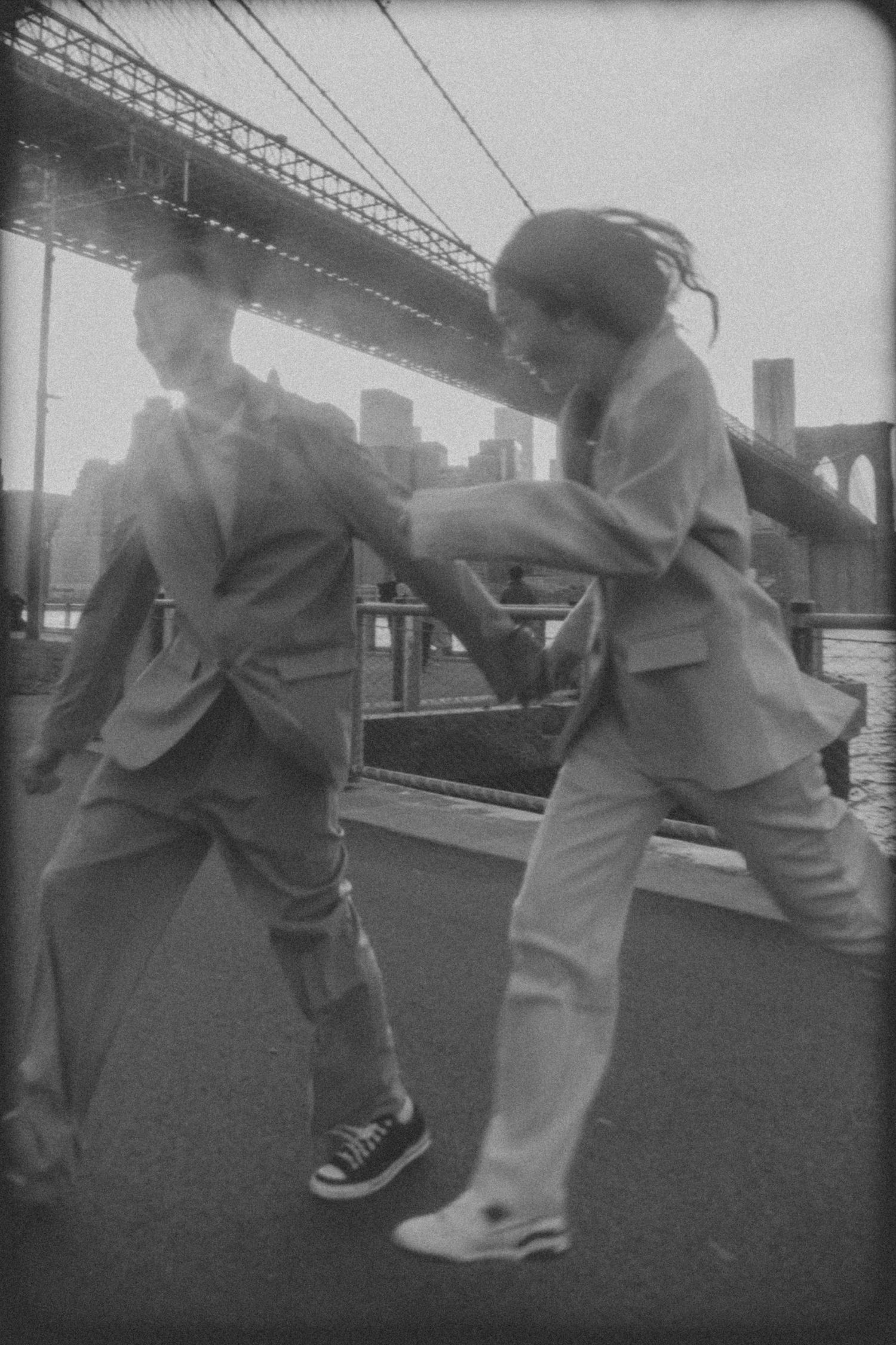 Two people dancing energetically in casual attire beneath the Brooklyn Bridge in a vintage black and white photograph.