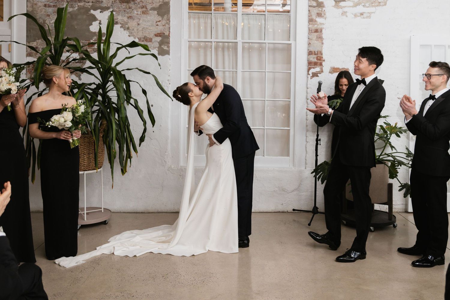 Wedding couple shares a kiss during their ceremony while guests and wedding party look on in an industrial chic venue.