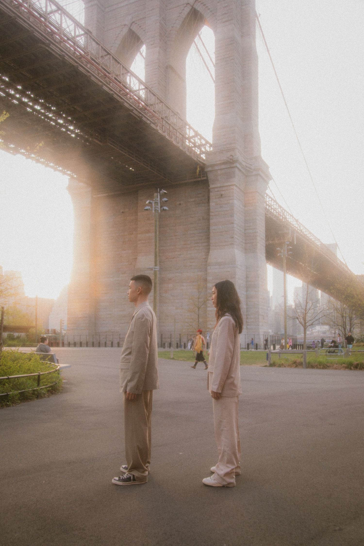 Two people in coordinated beige outfits standing beneath the majestic Brooklyn Bridge during golden hour.