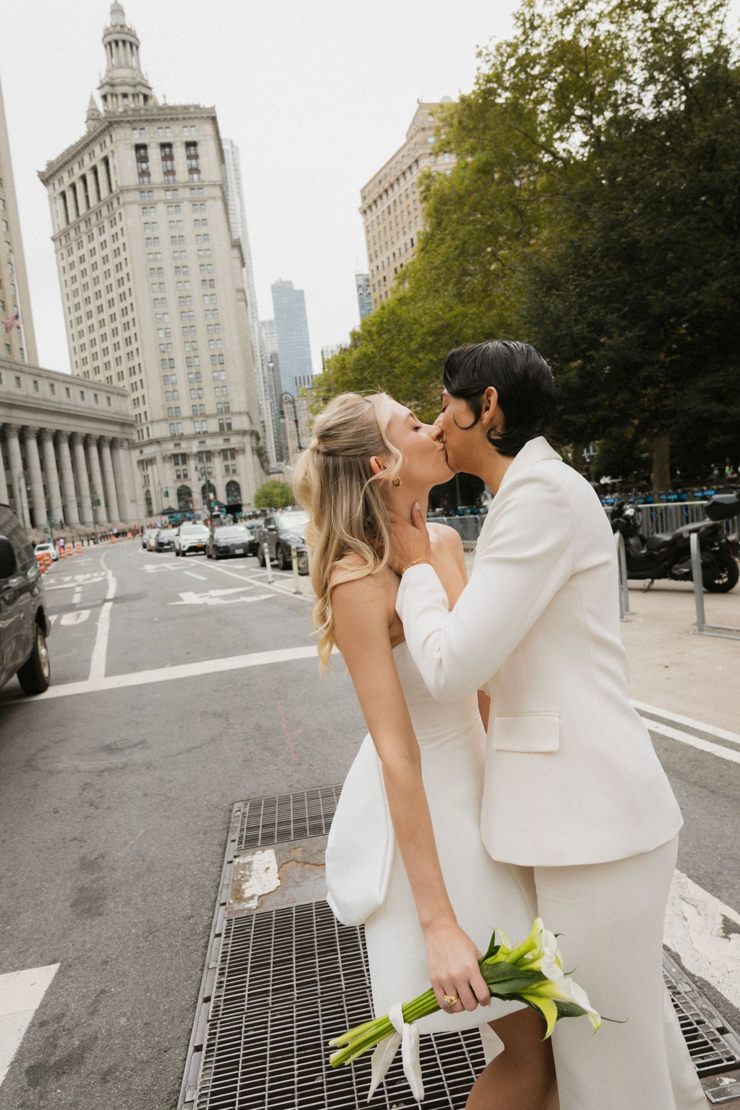 Newlywed couple shares romantic kisses on city street with urban architecture and buildings in background.