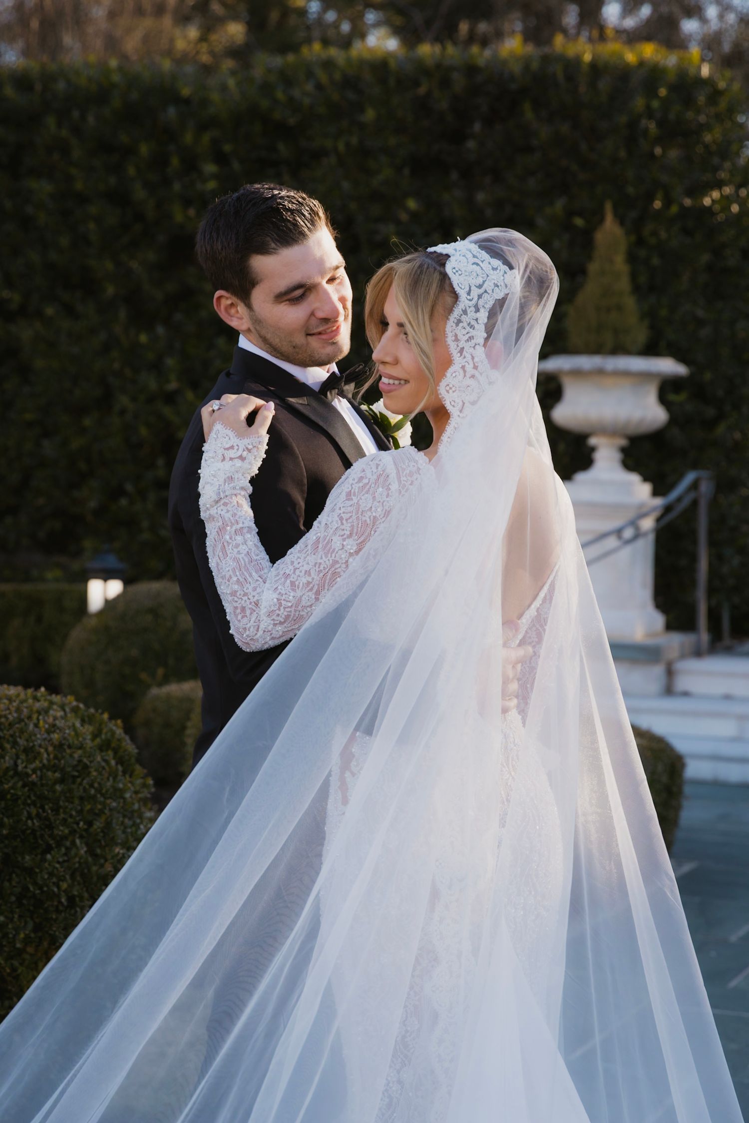 Newlywed couple shares romantic moments in elegant wedding attire with flowing white veil in garden setting.