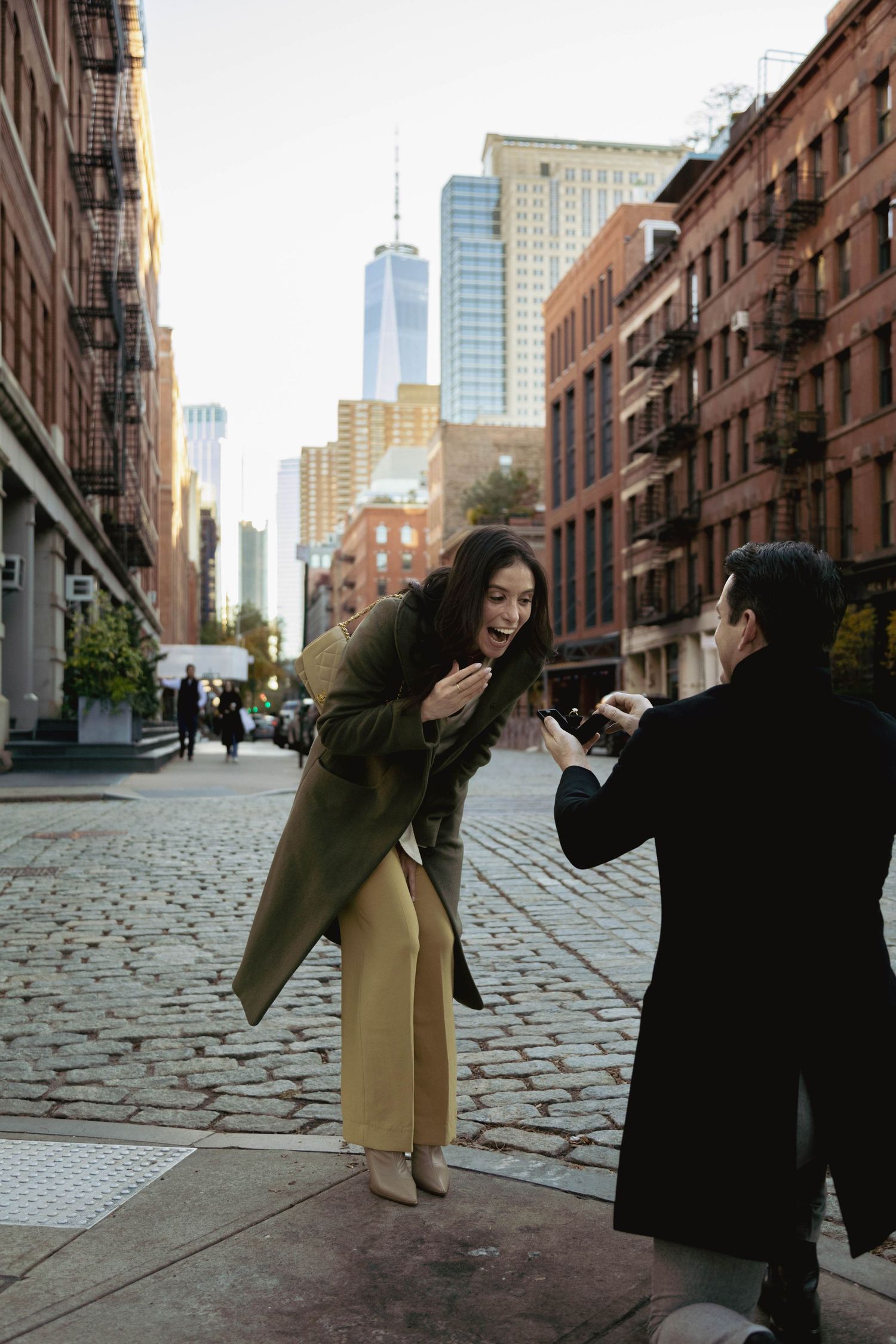 Two people in casual attire dance playfully on a cobblestone street in DUMBO Brooklyn with city buildings in background.