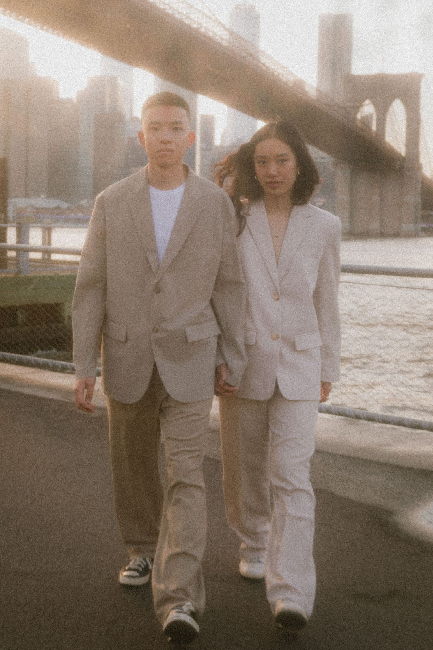 Two people in matching beige suits walk near the Brooklyn Bridge in New York City during golden hour.