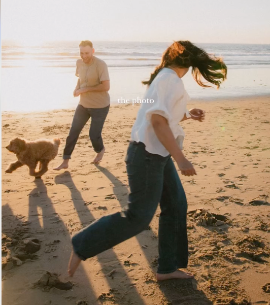 People dancing and running with a dog on a sunlit beach during golden hour.
