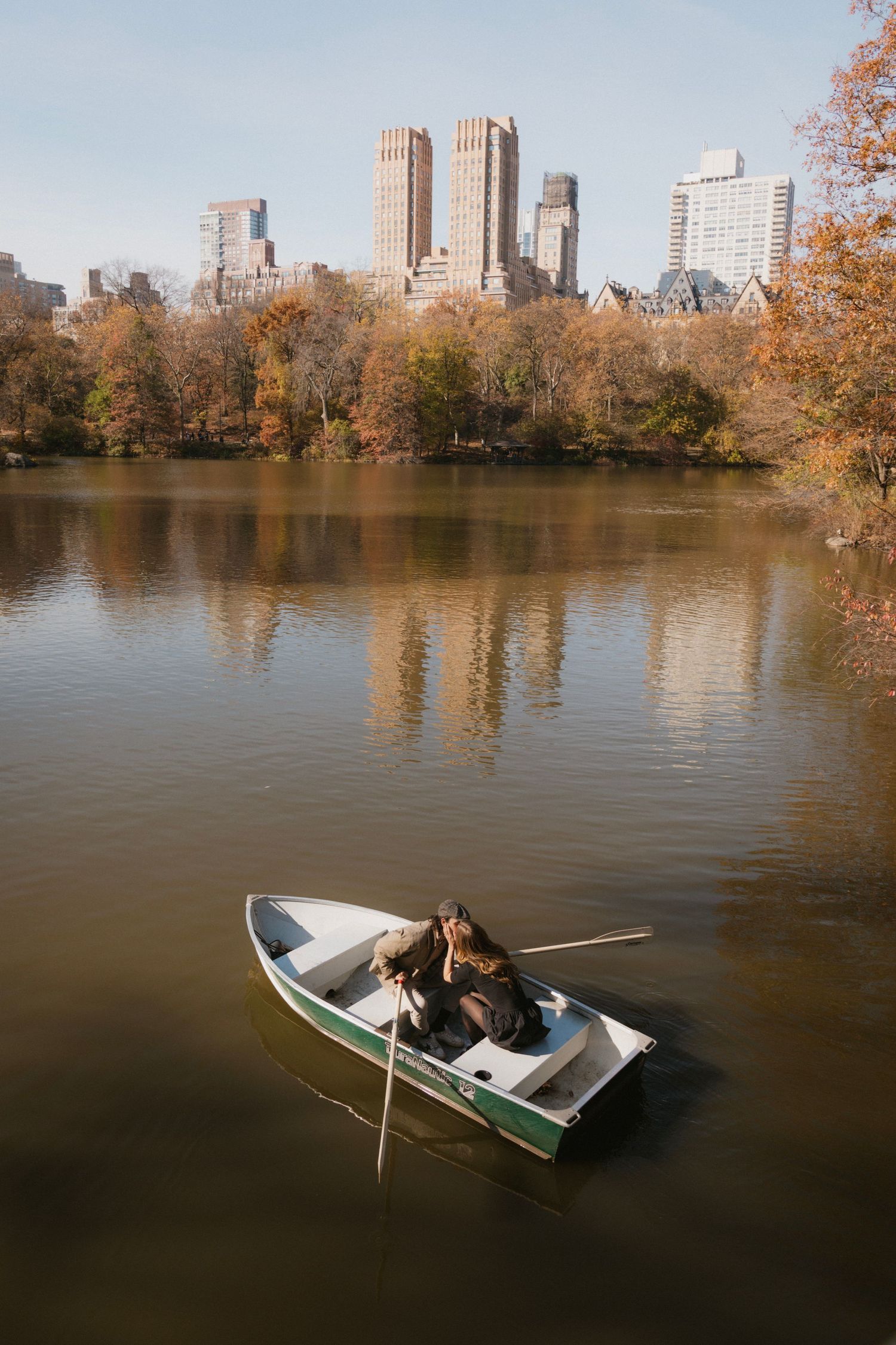 A romantic boat ride on a lake in Central Park during autumn with Manhattan skyscrapers reflecting in the calm water.